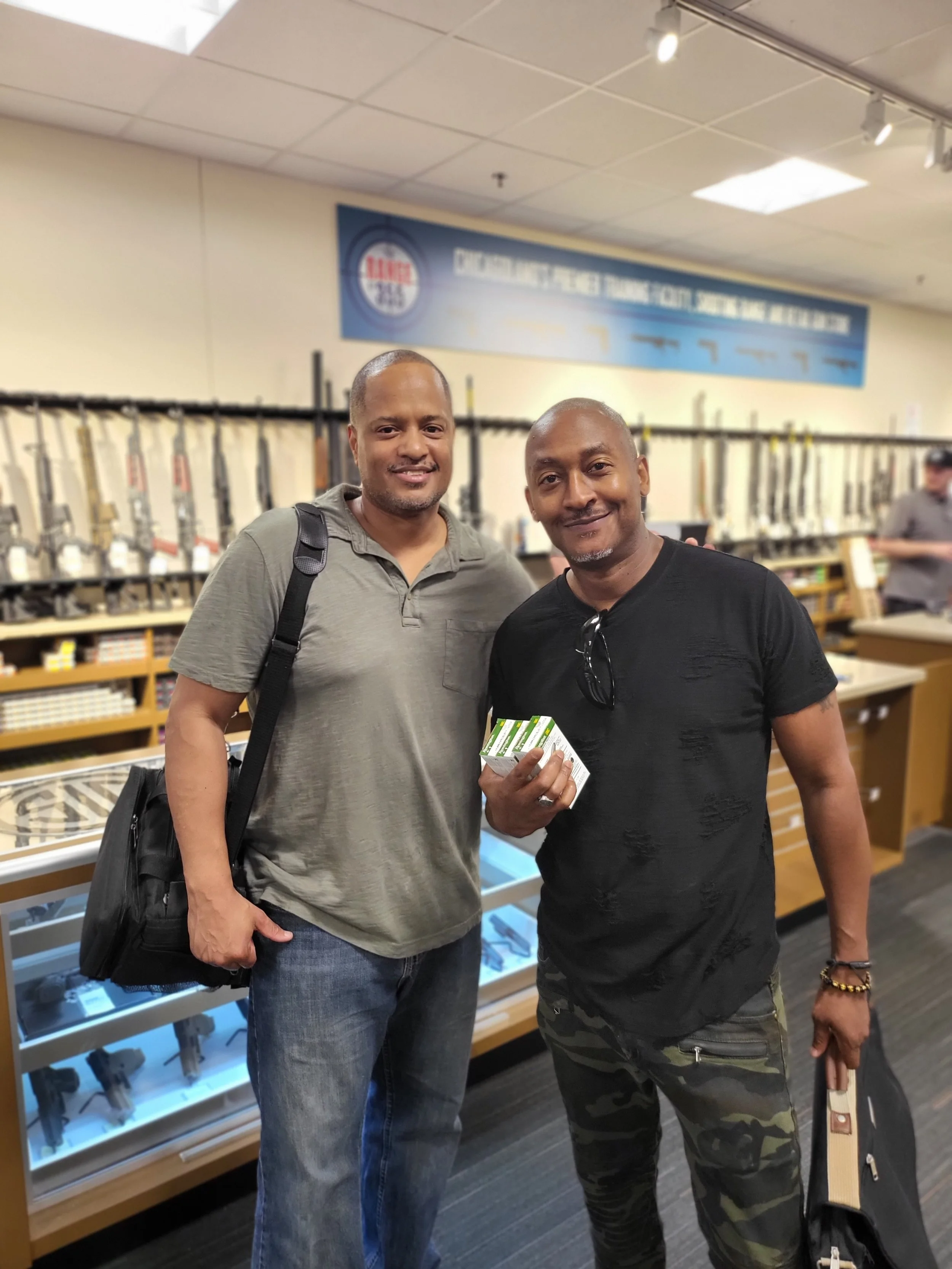 Two men standing inside a gun shop. One man is wearing a gray shirt and carrying a black bag, and the other is wearing a black shirt and camouflage pants, holding a box of ammunition. There are rifles displayed on the wall behind them.