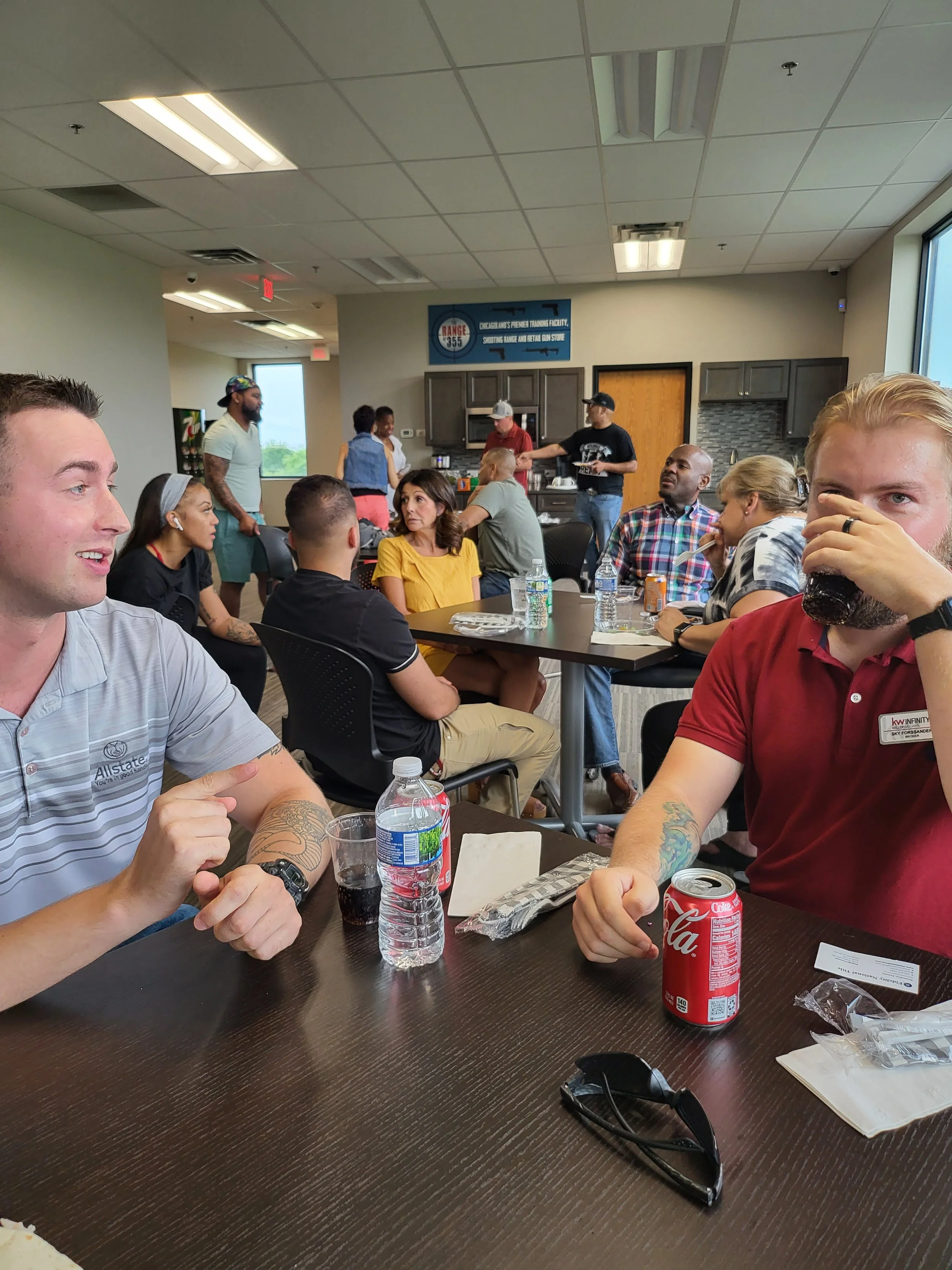 People sitting at a table in a room, some chatting, drinking soda, and eating, with a kitchen area in the background and a sign that says 'Range 355: Charcoalless Firearm Training Facility, Shooting Range and Retail Gun Store' on the wall.