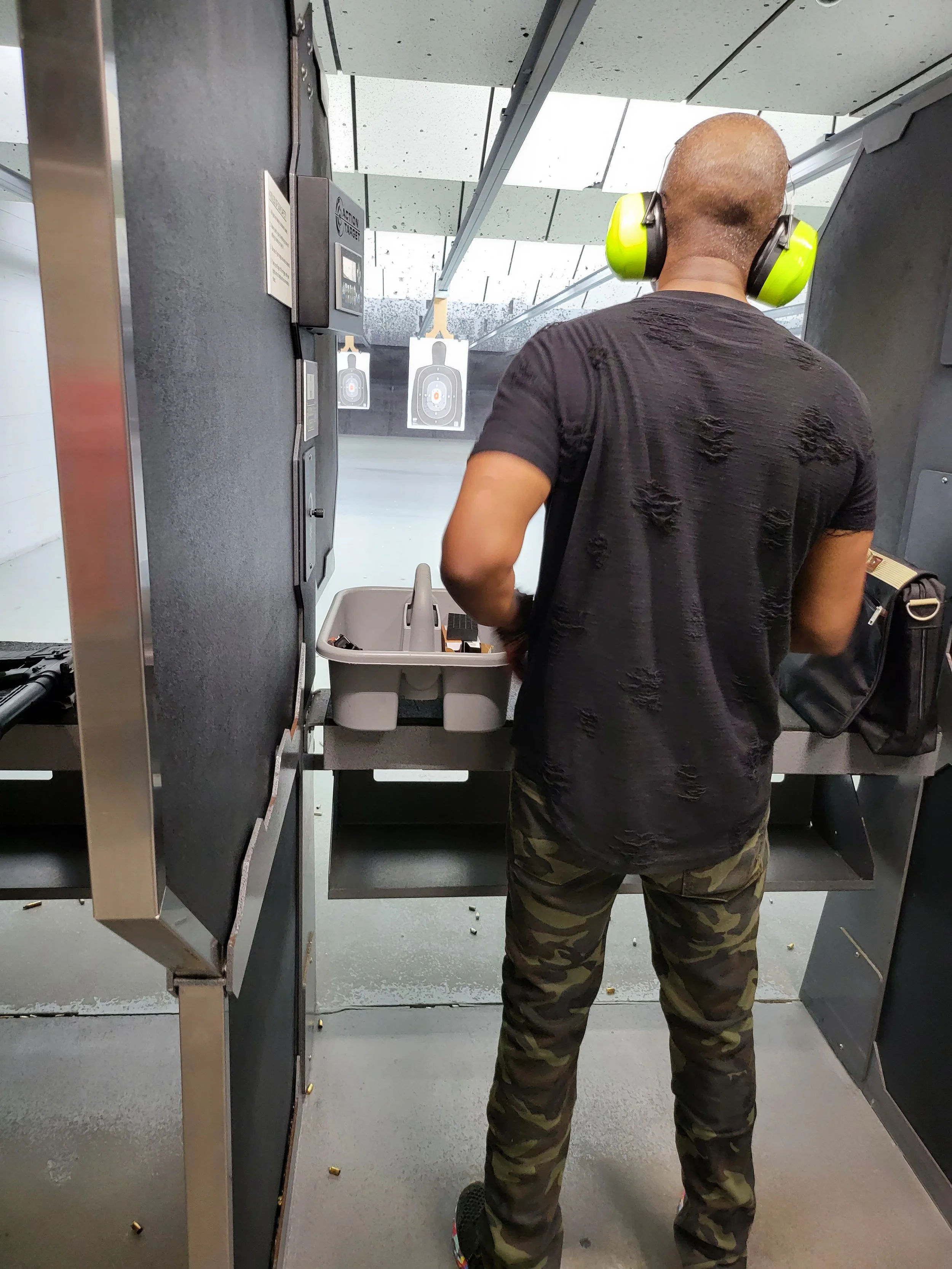 A person with a shaved head wearing yellow ear protectors, a black distressed shirt, and camo pants, at a shooting range, aiming at targets in the distance.