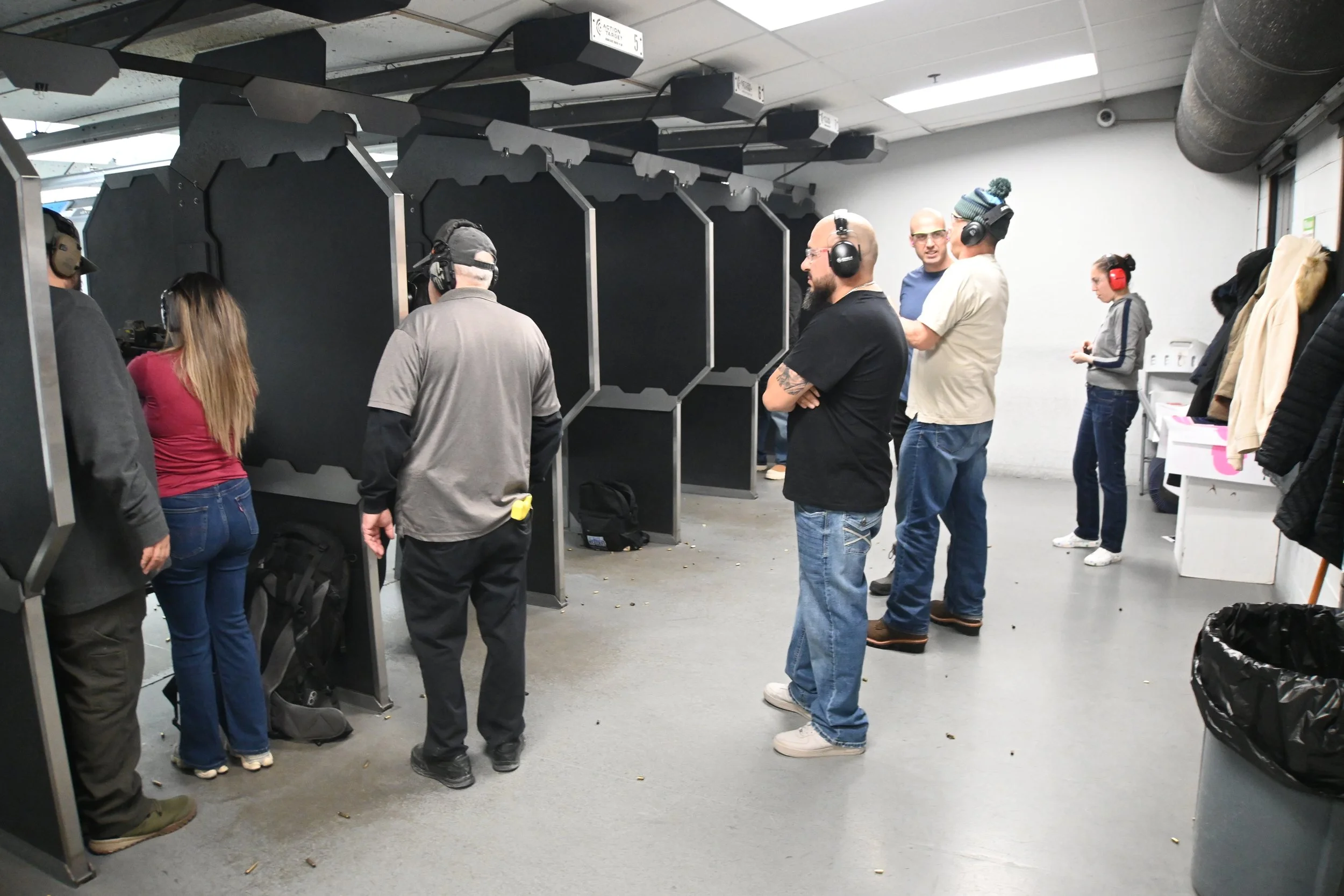 People standing and talking in a shooting range with black partitions and some people wearing ear protection.