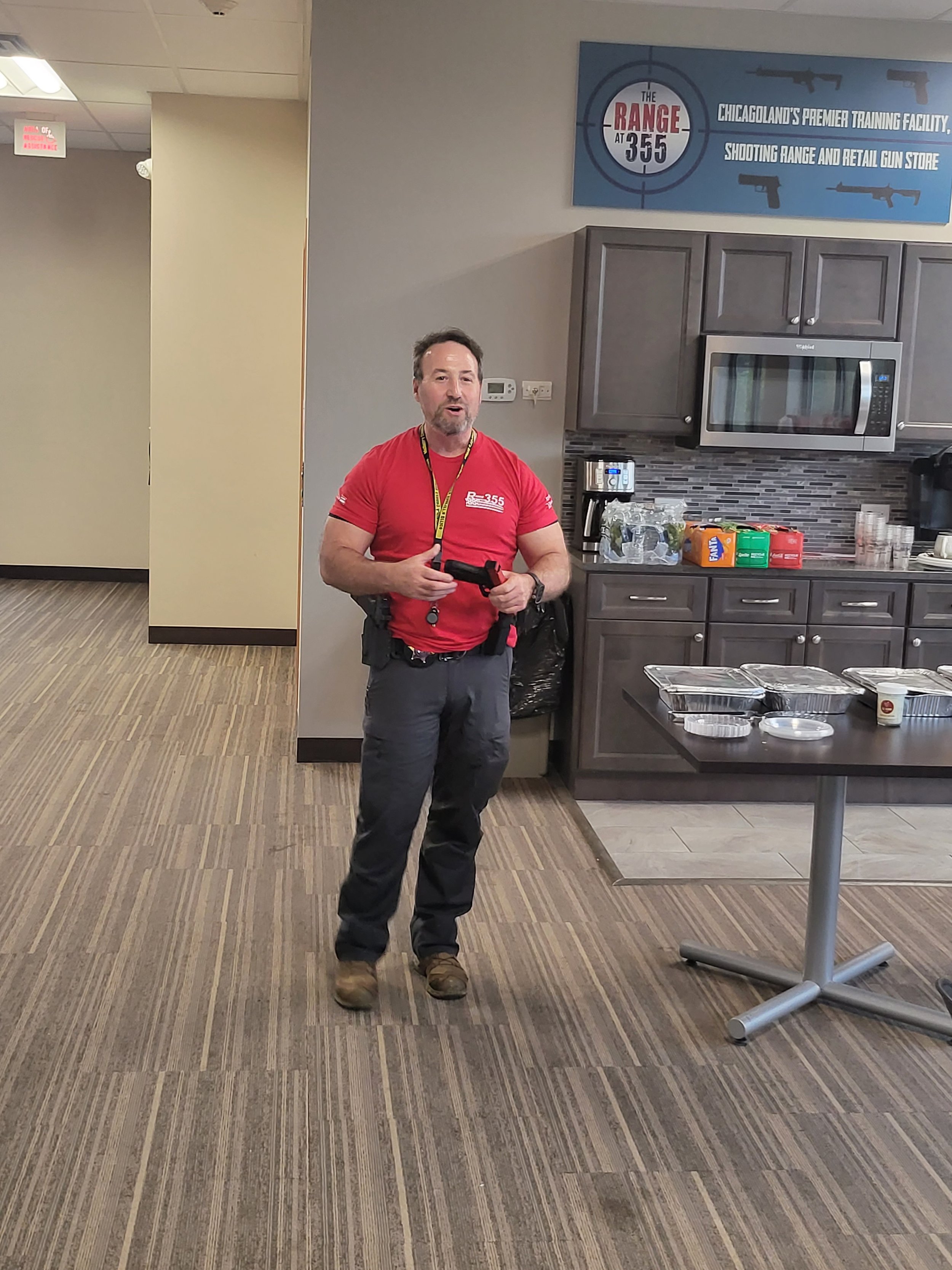 A man wearing a red shirt with a range logo, holding a gun, standing in a breakroom with a table, microwave, and snack items.