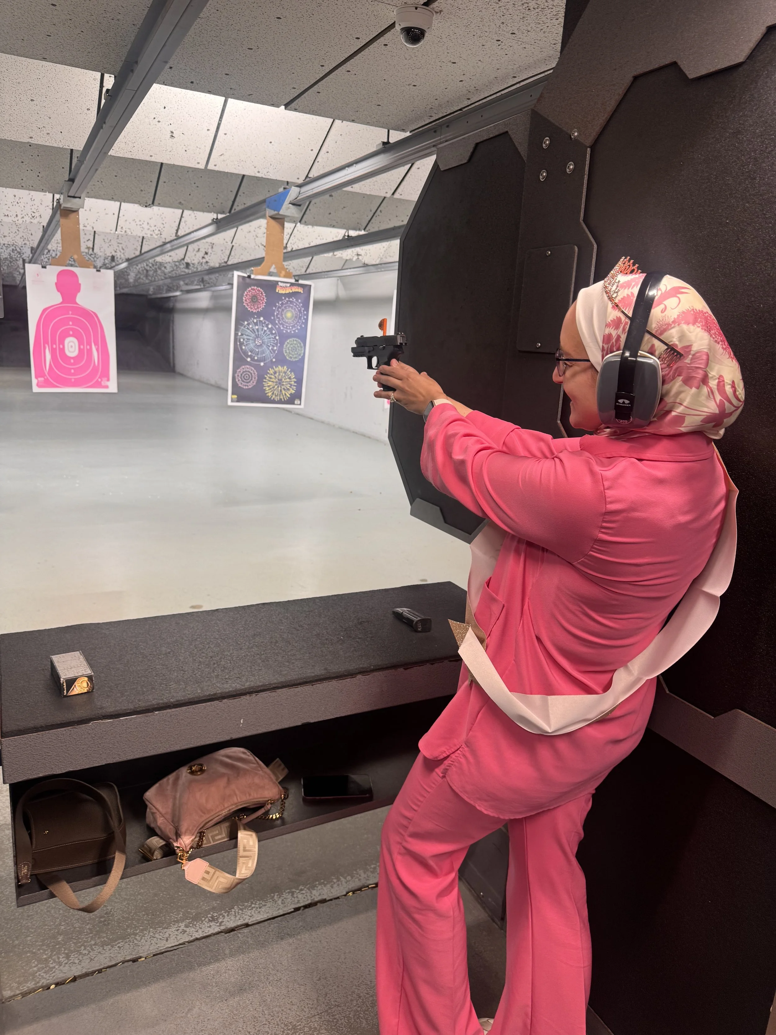 A woman wearing pink clothing and a floral headscarf is shooting a handgun at an indoor shooting range. She is wearing ear protection and glasses, and is aiming at targets down the range.