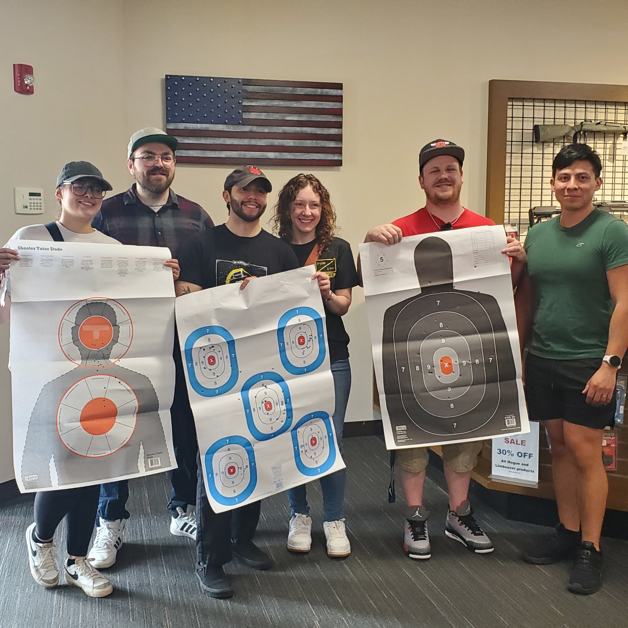 A group of six people standing indoors, holding target shooting posters, with a framed American flag on the wall behind them.