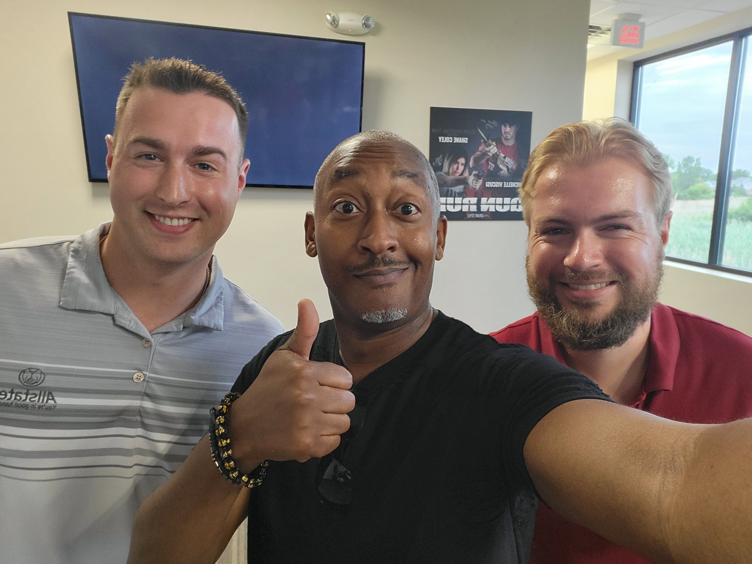 Three smiling men taking a selfie in an indoor setting with posters and windows in the background.