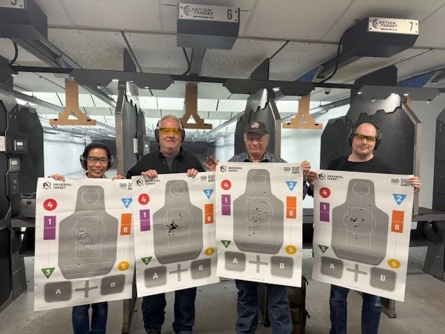 Four adults at an indoor shooting range holding up target sheets with bullet holes, wearing eye protection, behind shooting booths.