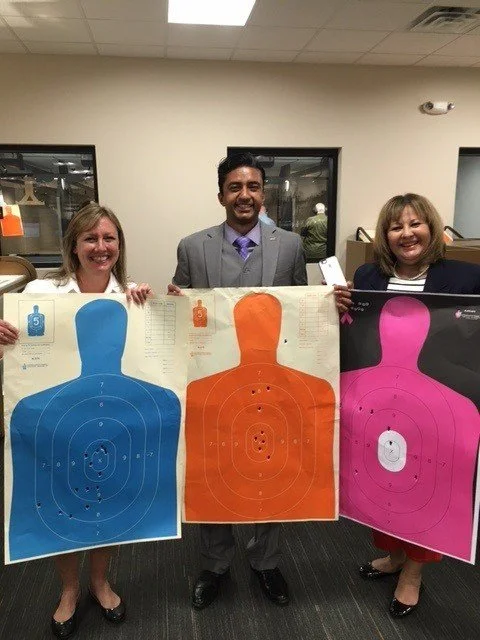 Three people standing indoors, each holding a paper shooting range target. The targets are colored blue, orange, and pink, each with concentric circles and a silhouette shape at the top.