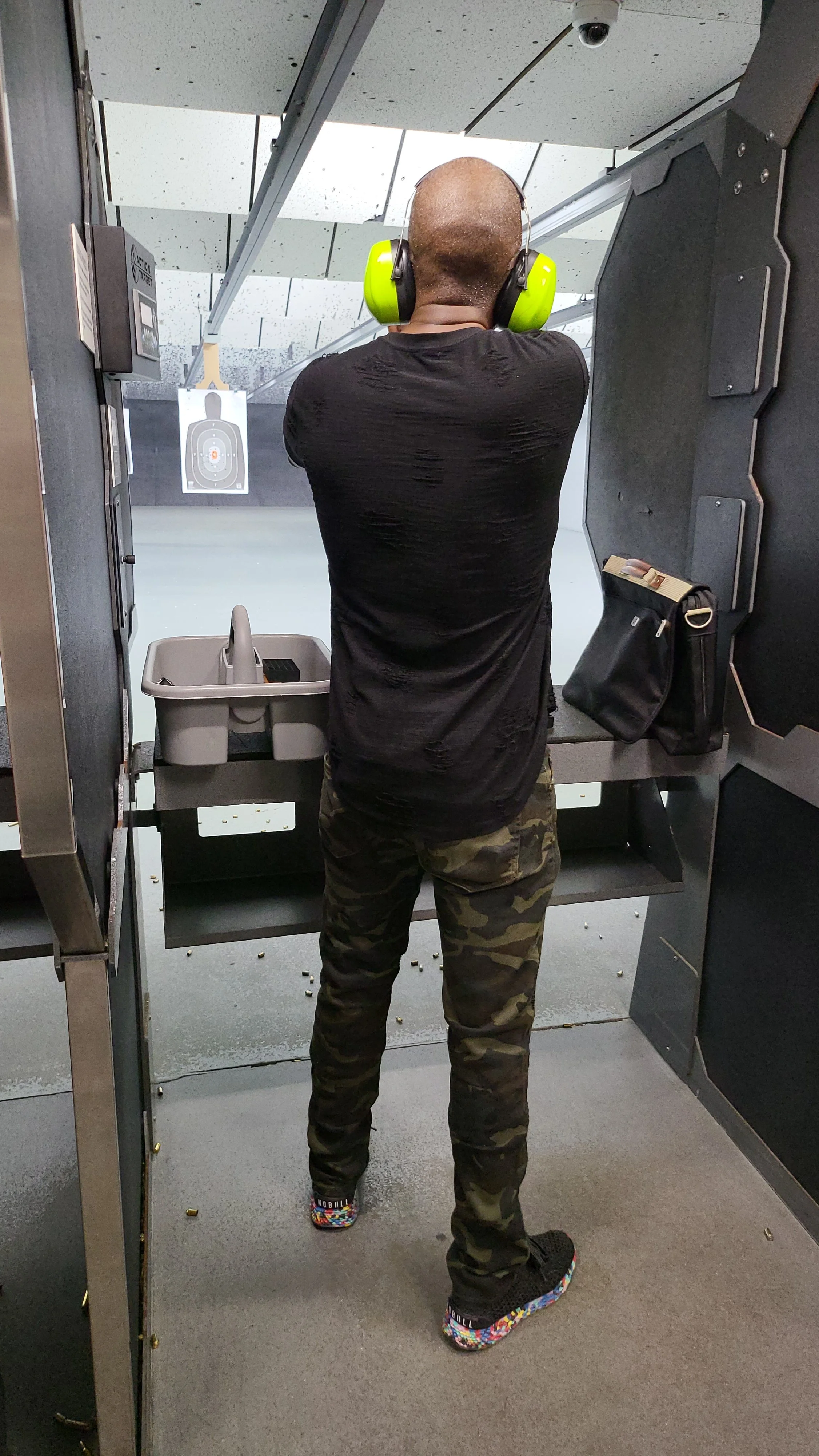A person at an indoor shooting range, wearing green ear protection and camouflage pants, standing at a shooting station facing a target in the distance.