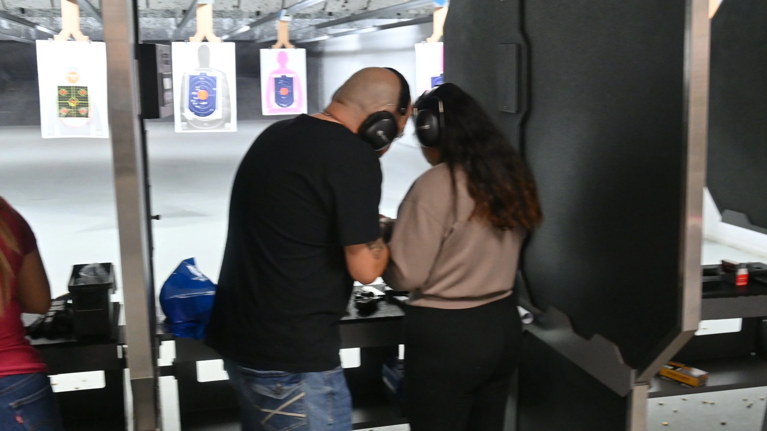 Two people are at an indoor shooting range, wearing hearing protection, and appear to be examining or discussing something on the table in front of them. There are target sheets hanging in the background.