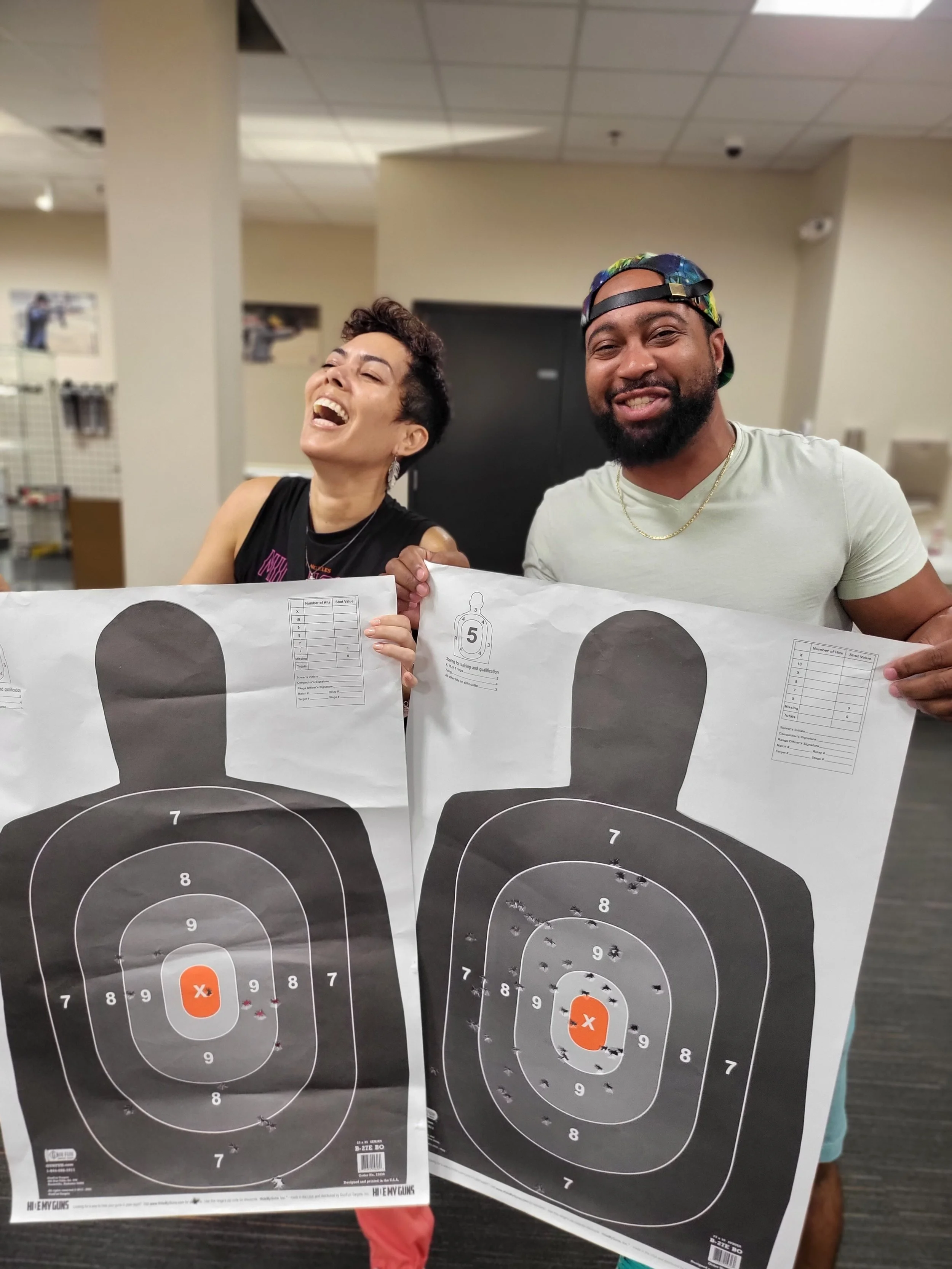 Two people standing behind shooting targets at an indoor shooting range, holding paper targets with bullet holes, one woman laughing and a man smiling.
