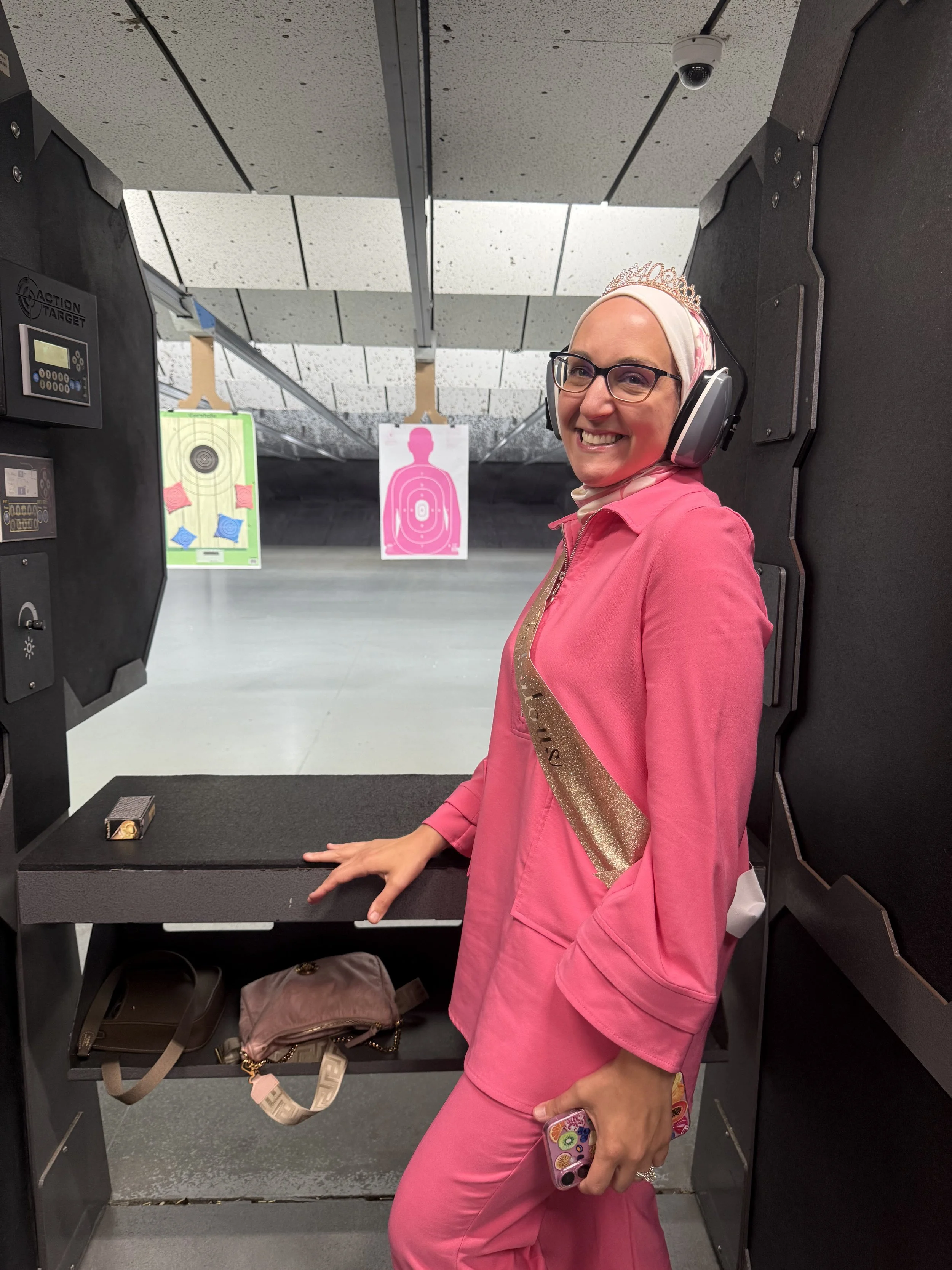 A woman wearing pink scrubs, glasses, a tiara, and a sash labeled 'birthday girl' at a shooting range, smiling and holding a phone with colorful case.