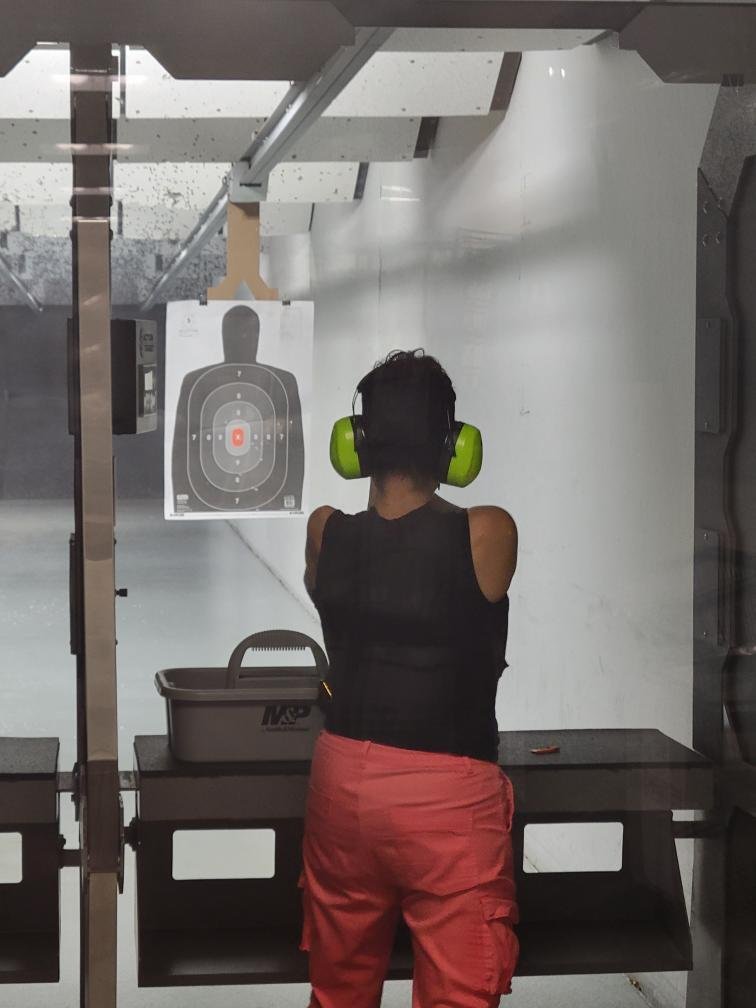 Person wearing green ear protectors at an indoor shooting range, aiming at a paper target with a silhouette and scoring rings, standing behind a table with a black container.