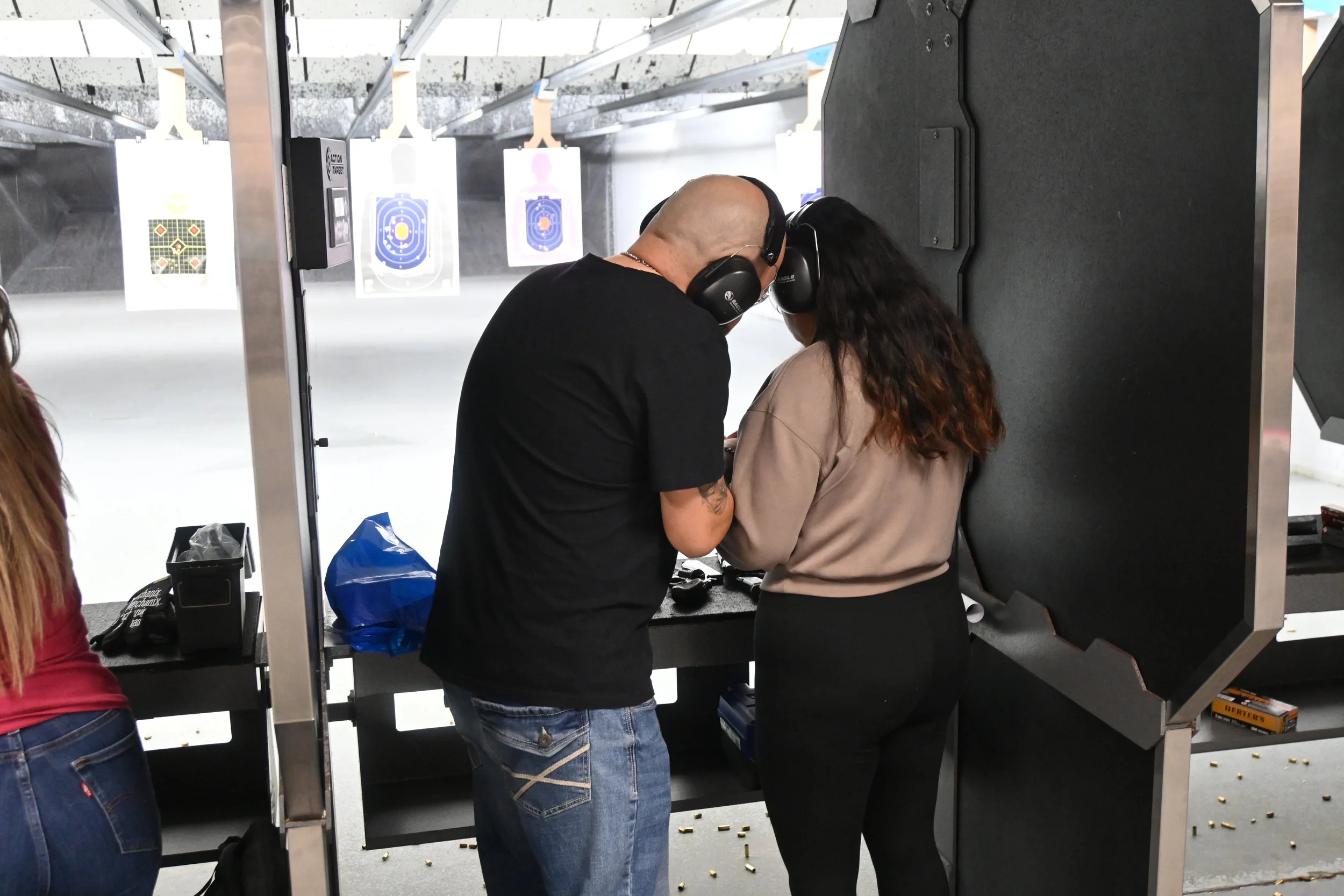 Two people at an indoor shooting range wearing hearing protection, standing close together, possibly inspecting or discussing something. Target papers are hanging in the background.