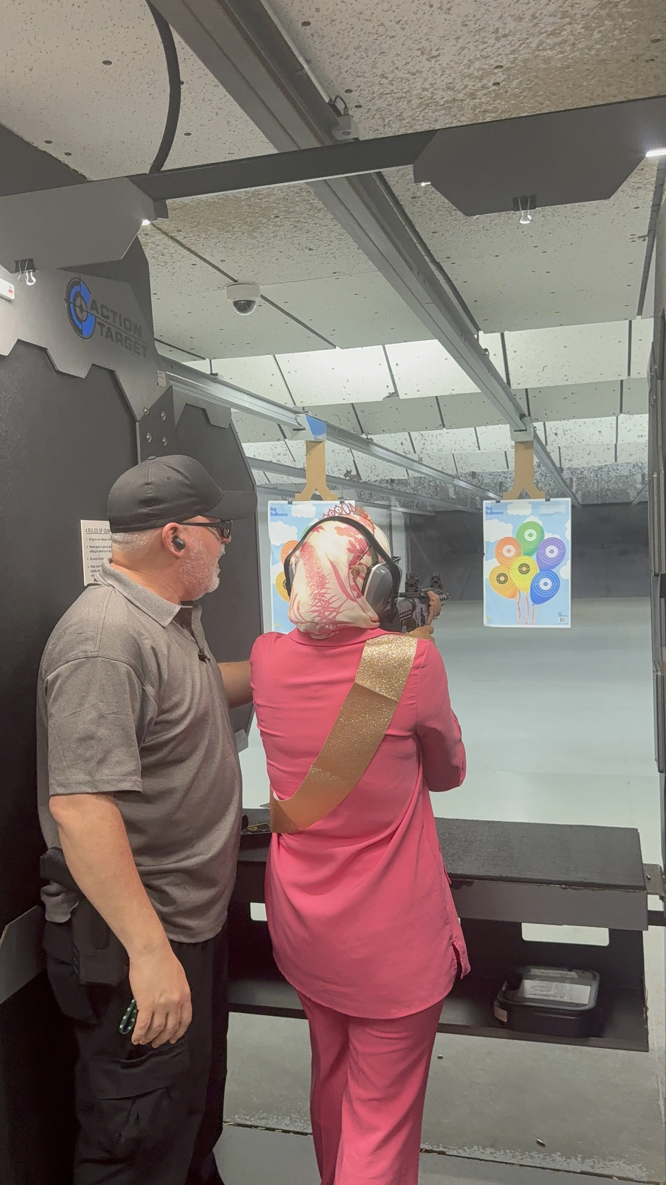 Two people, a man and a woman, are at a shooting range. The woman is aiming a rifle at targets downrange, and the man is standing beside her. The woman is wearing a pink outfit with a gold sash, and the man is wearing a gray shirt, a black cap, and g