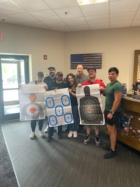 Group of six people holding target sheets in an indoor space with an American flag on the wall.