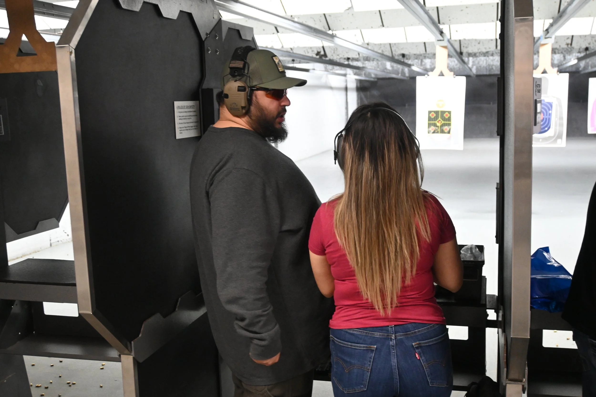Two people at a shooting range, one man wearing a cap, headphones, and sunglasses, and a woman with long hair and a red shirt facing target sheets with shooting targets.