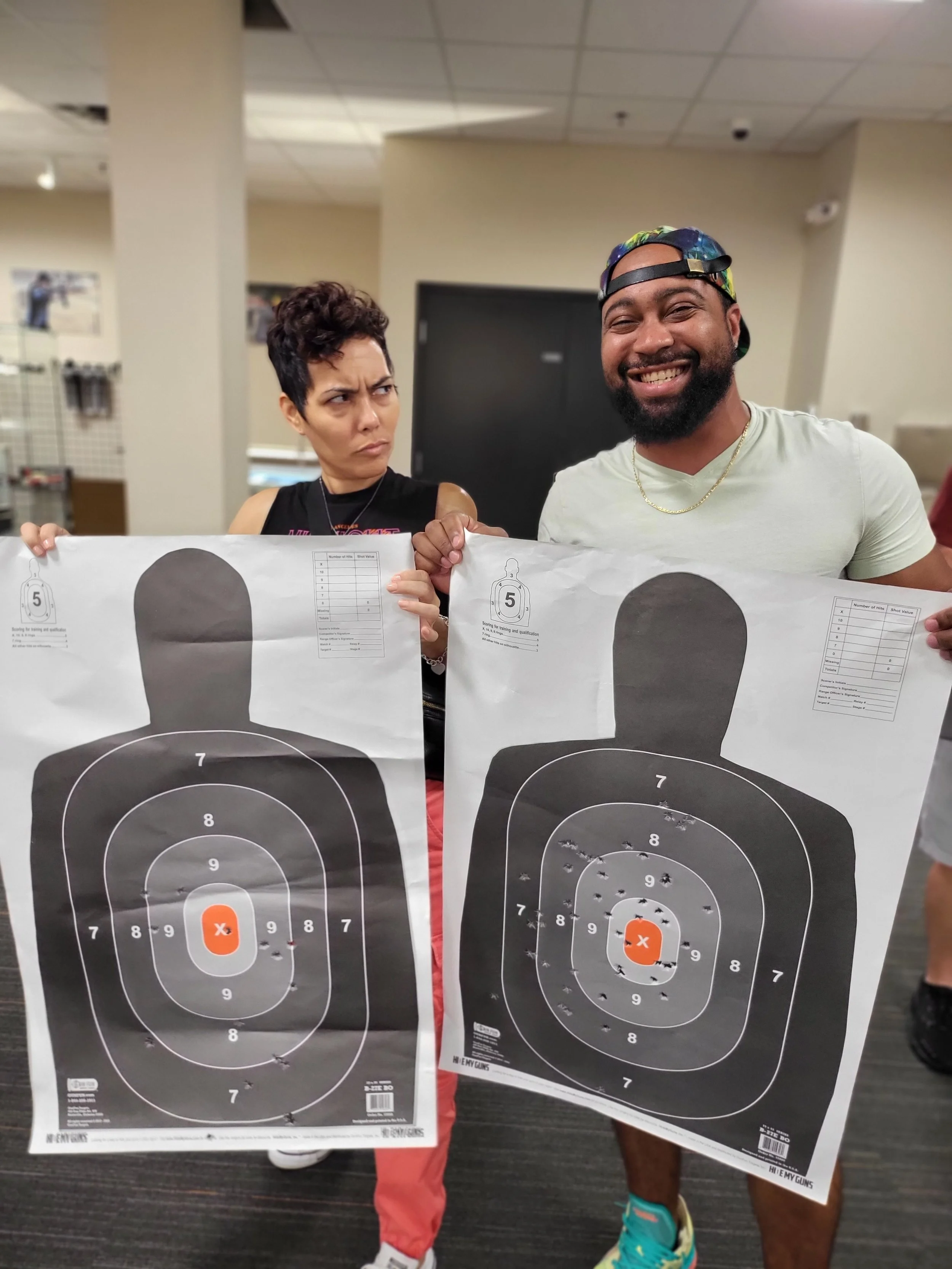 Two people holding shooting targets with bullet holes, one person is giving a confused look while the other is smiling.