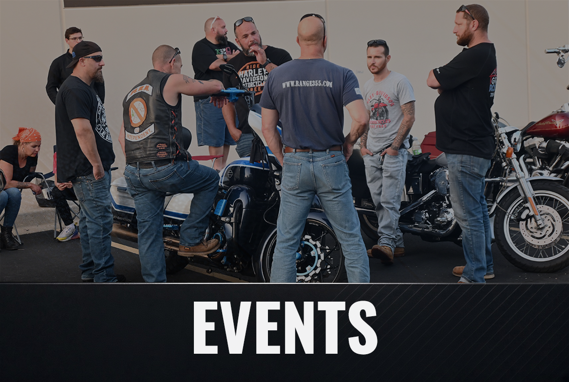 Group of people gathered around a motorcycle at a motorcycle event, some wearing Harley Davidson t-shirts and accessories, with a sign that says 'EVENTS' at the bottom.