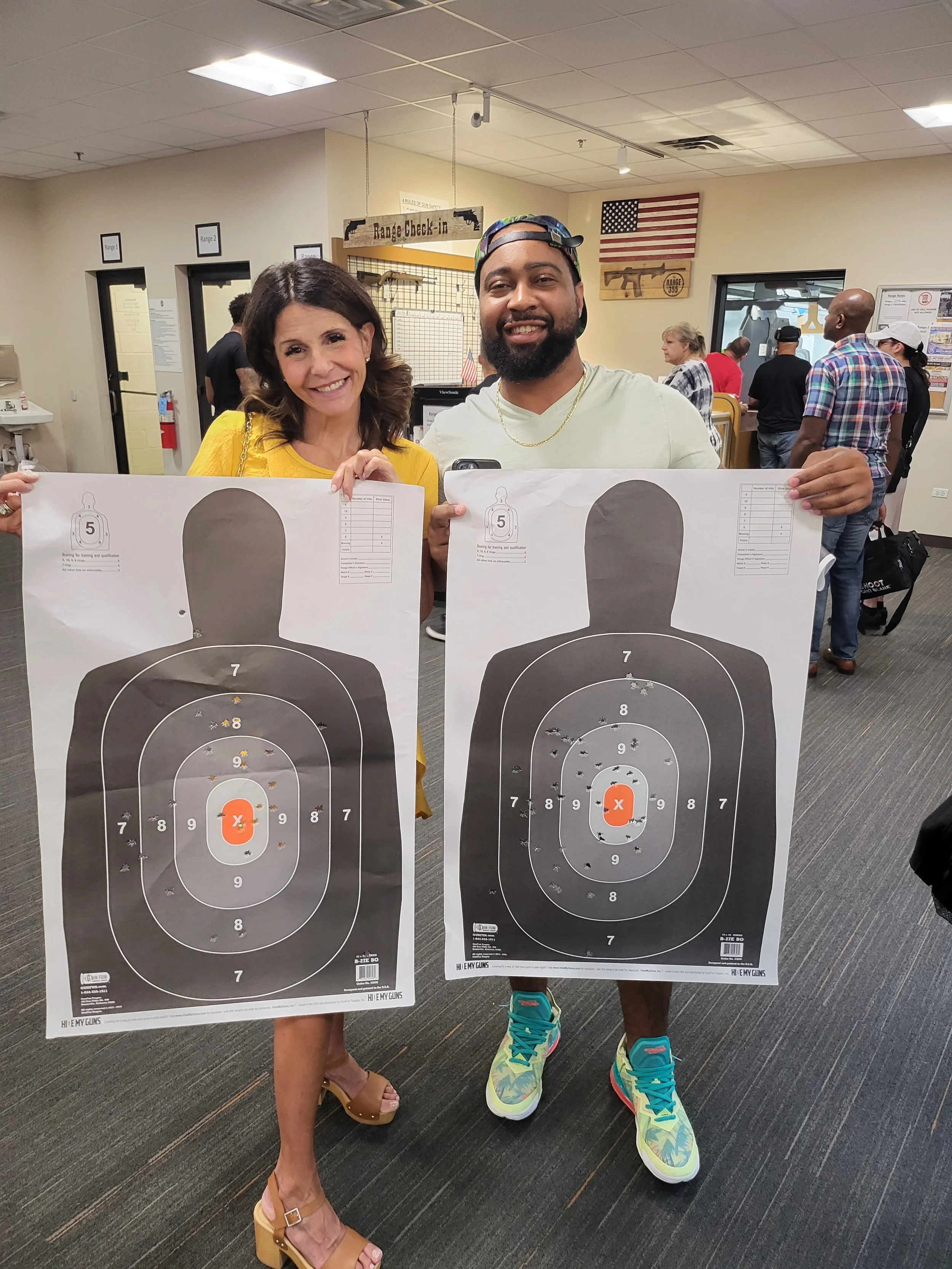 Two individuals holding target sheets at a shooting range. The targets have number scores and one shot marked with an orange X. The background shows a busy indoor shooting range with people and American flags.