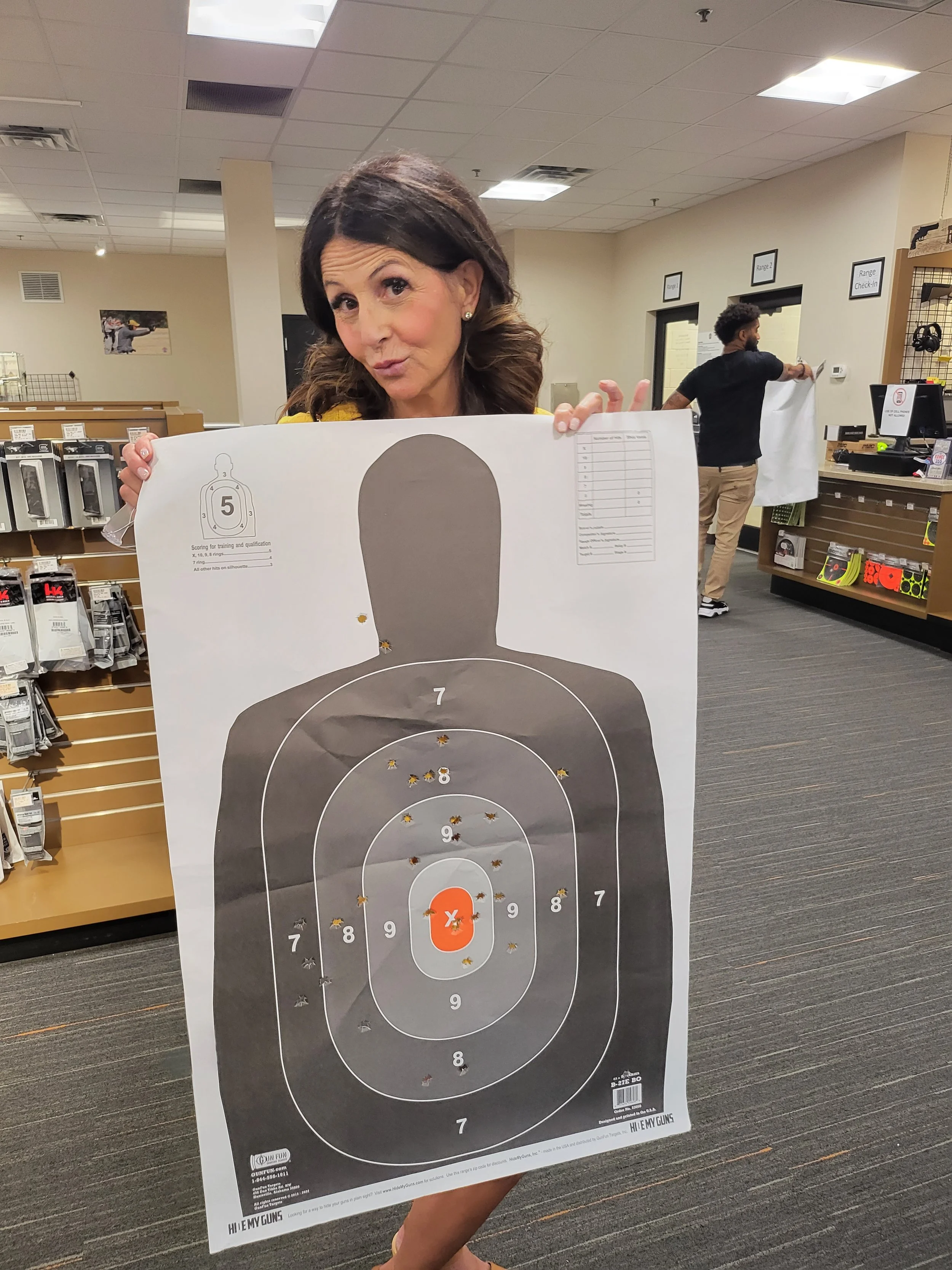 A woman is holding a shooting target sheet with a large black silhouette and concentric rings numbered 7 to 10, with bullet holes marked on the sheet, in an indoor shooting range.