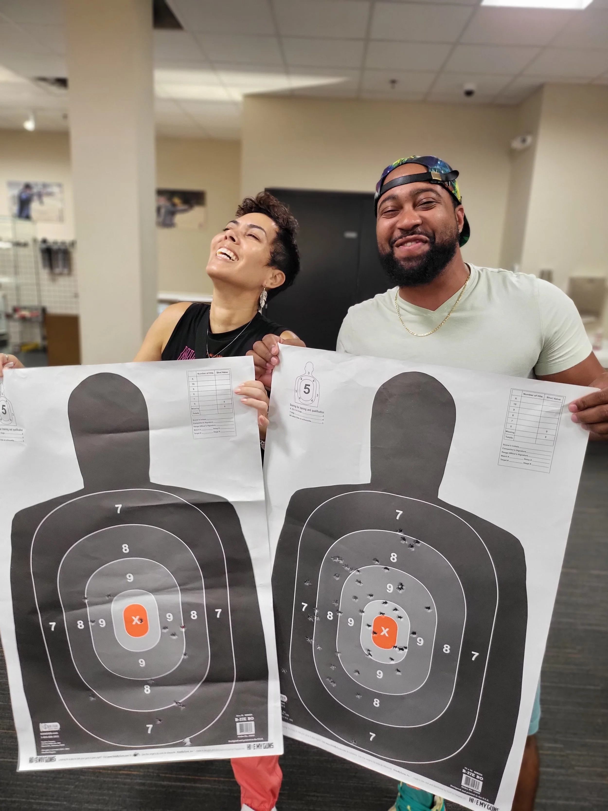 Two people holding up target sheets at an indoor shooting range, with a woman smiling and a man with a beard smiling, both showing the targets with bullet holes.