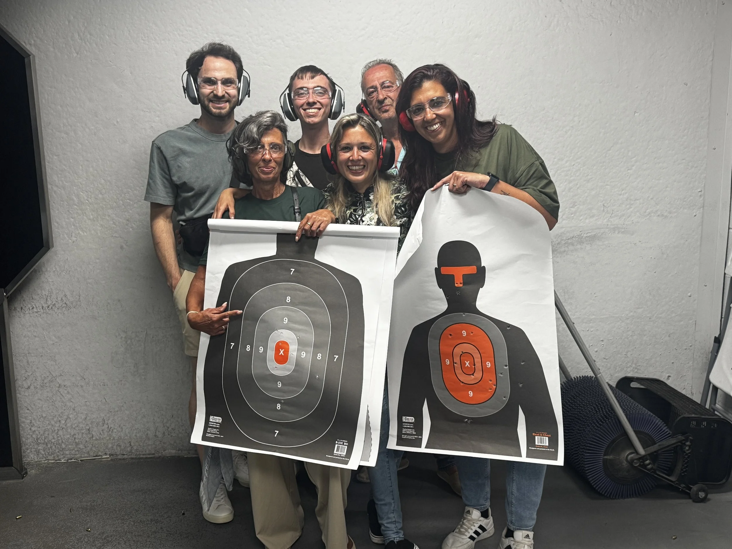 A group of seven people at a shooting range holding target sheets, some wearing protective ear and eye gear, smiling.