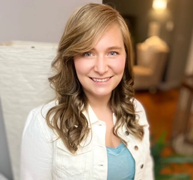 A headshot of Jessica Brewster, RMT. Jessica is a white woman with curly light brown hair and light eyes. She is wearing a blue v-neck tee and white denim jacket.