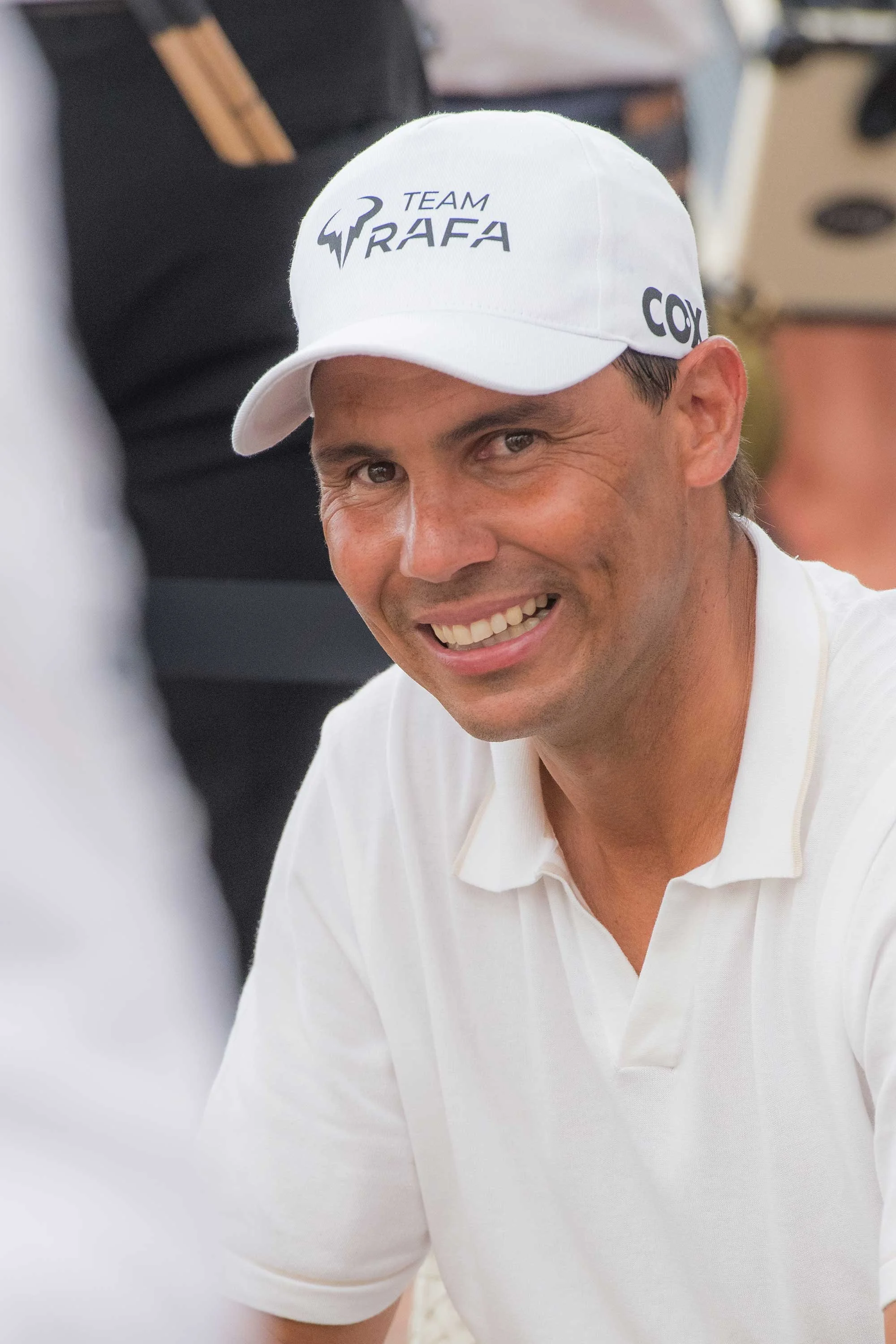 A man smiling, wearing a white cap with the logo 'Team Rafa.' He appears to be outdoors or at a sporting event.