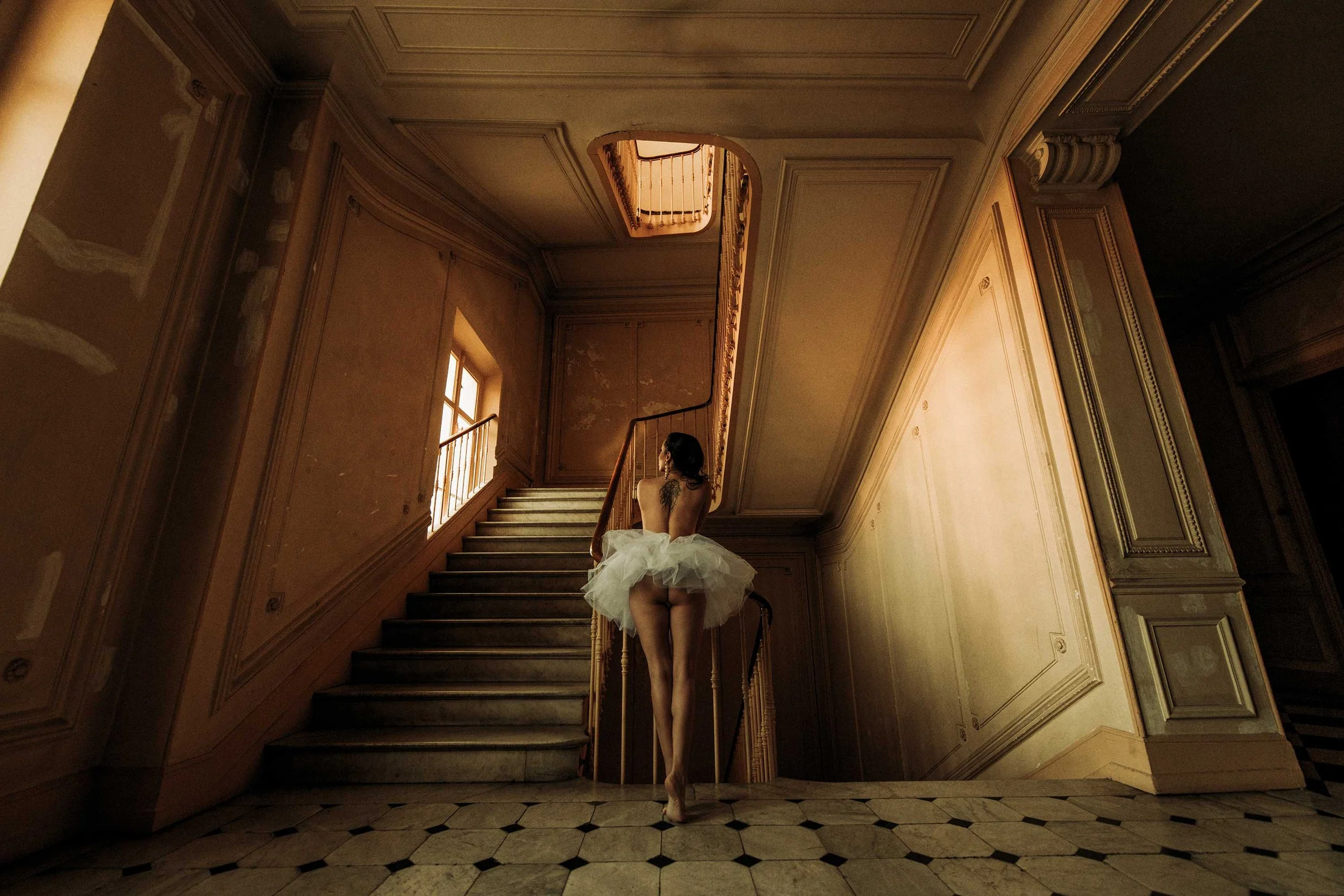 A woman wearing a white tutu standing on her toes at the bottom of a staircase in an ornate indoor setting, with light coming through windows.