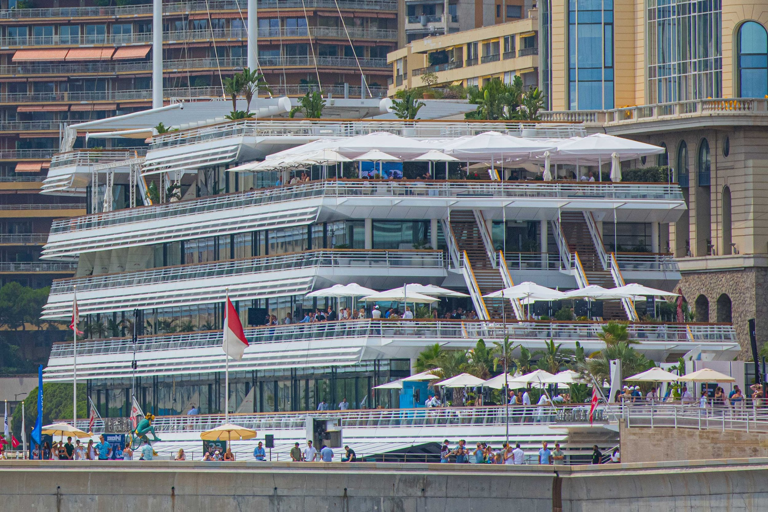 Multi-level modern building with outdoor dining areas, white umbrellas, and people dining, located near water with flags and palm trees.