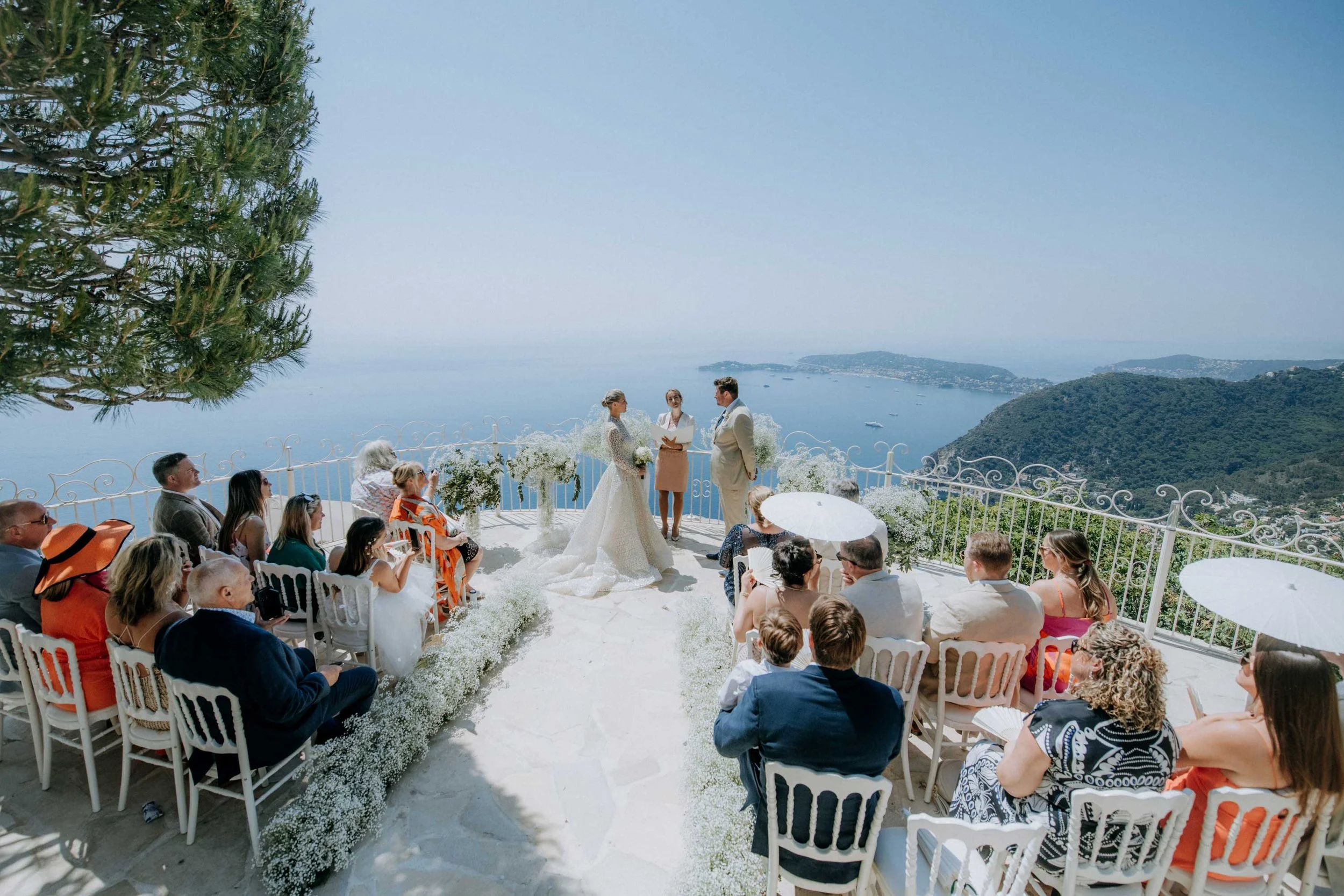 A wedding ceremony taking place outdoors on a terrace overlooking the ocean and hills, with the bride and groom standing facing each other and an officiant. Guests are seated on either side, some under umbrellas, with a scenic coastal background.