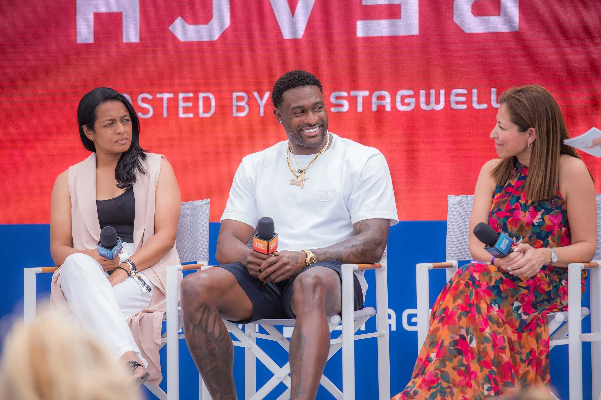 Three people sitting on stage during an event, each holding a microphone. The background displays a red and blue backdrop with text.