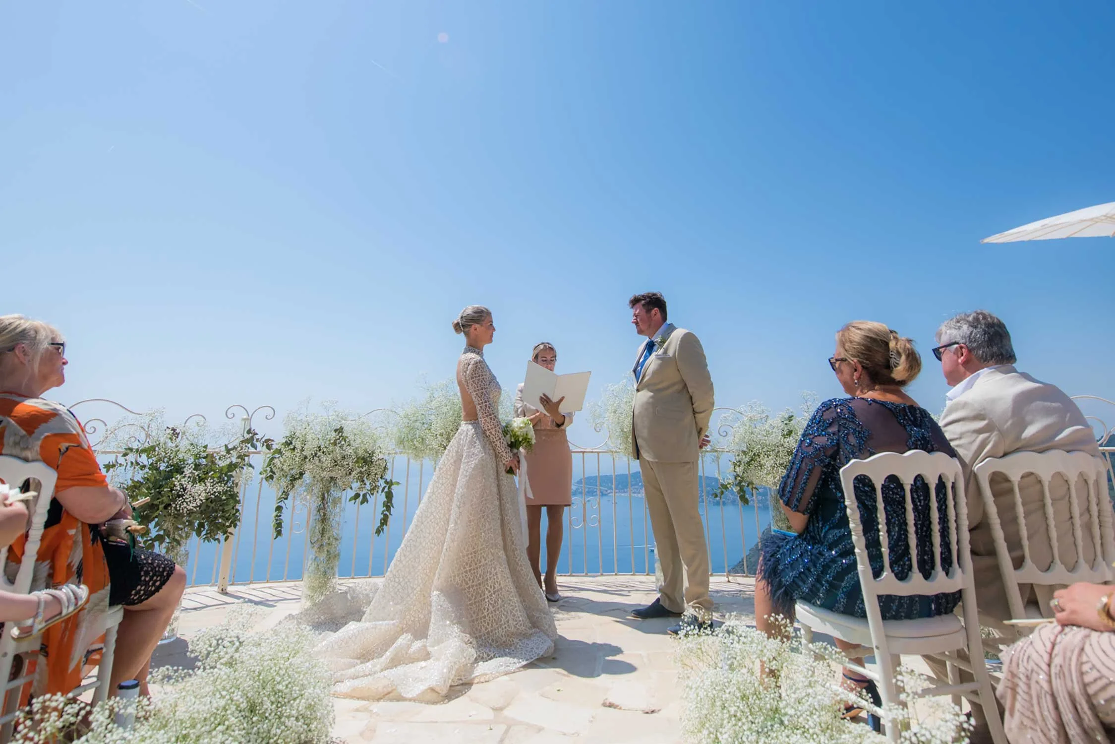 A wedding ceremony taking place outdoors on a scenic hill with a view of water and mountains in the background. The bride and groom are standing at the altar facing each other, with the officiant reading from a book. Guests are seated on white chairs decorated with flowers, and the sky is clear and blue.