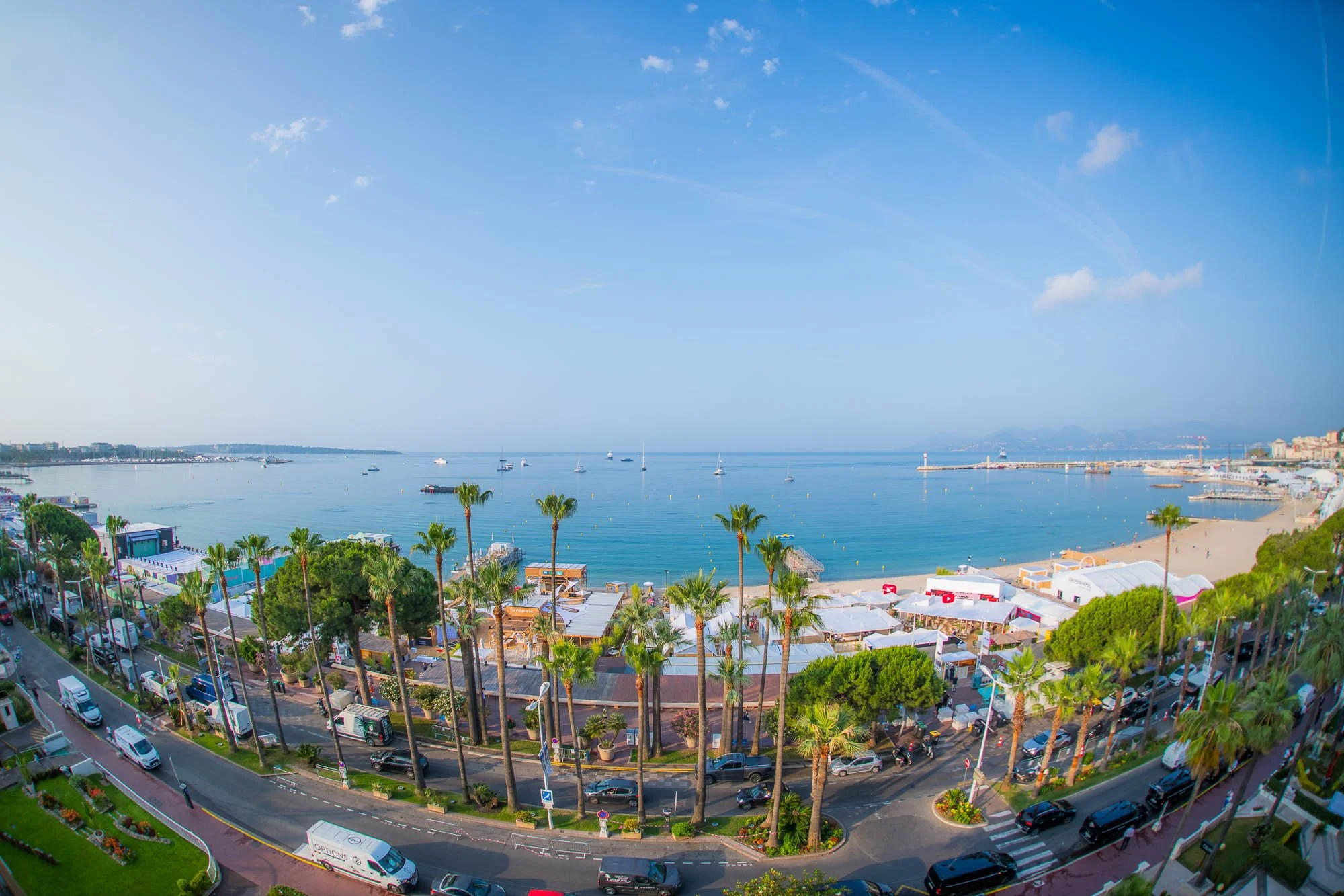 A scenic view of a coastal city with palm trees, a busy street, boats in the water, sandy beach, and a clear blue sky.