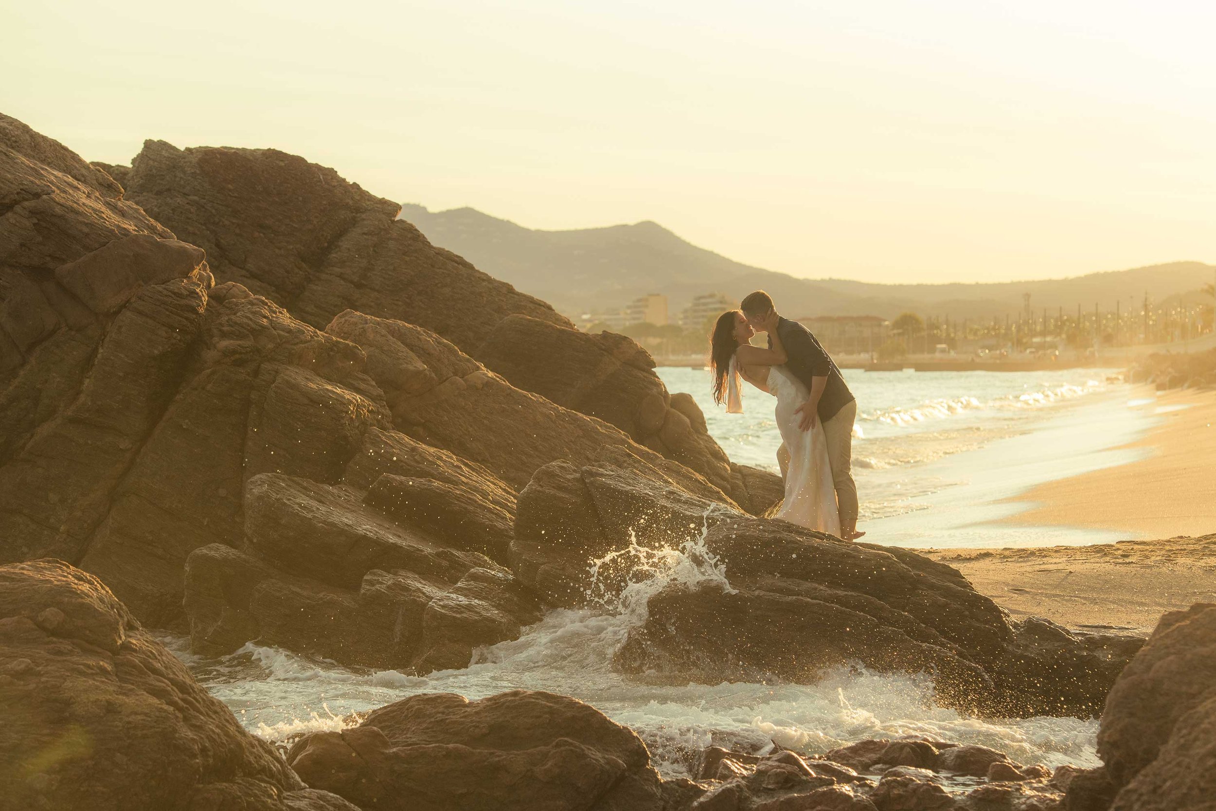 A couple in wedding attire kissing on a rocky beach at sunset, with the ocean waves crashing around them.