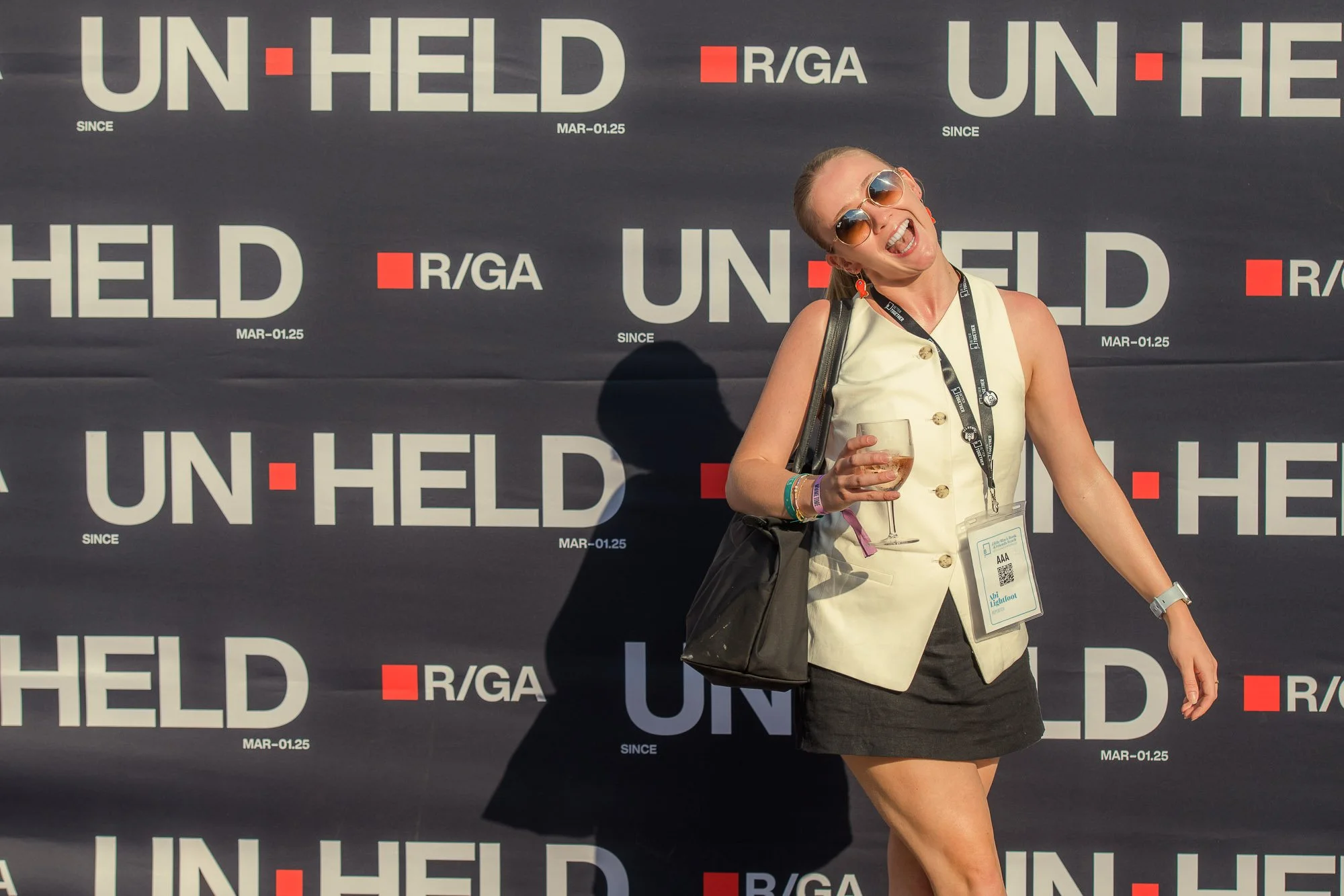 A smiling woman wearing sunglasses, holding a glass of wine, standing in front of a backdrop that reads 'UN-HLED' and 'R/GA' during an event.