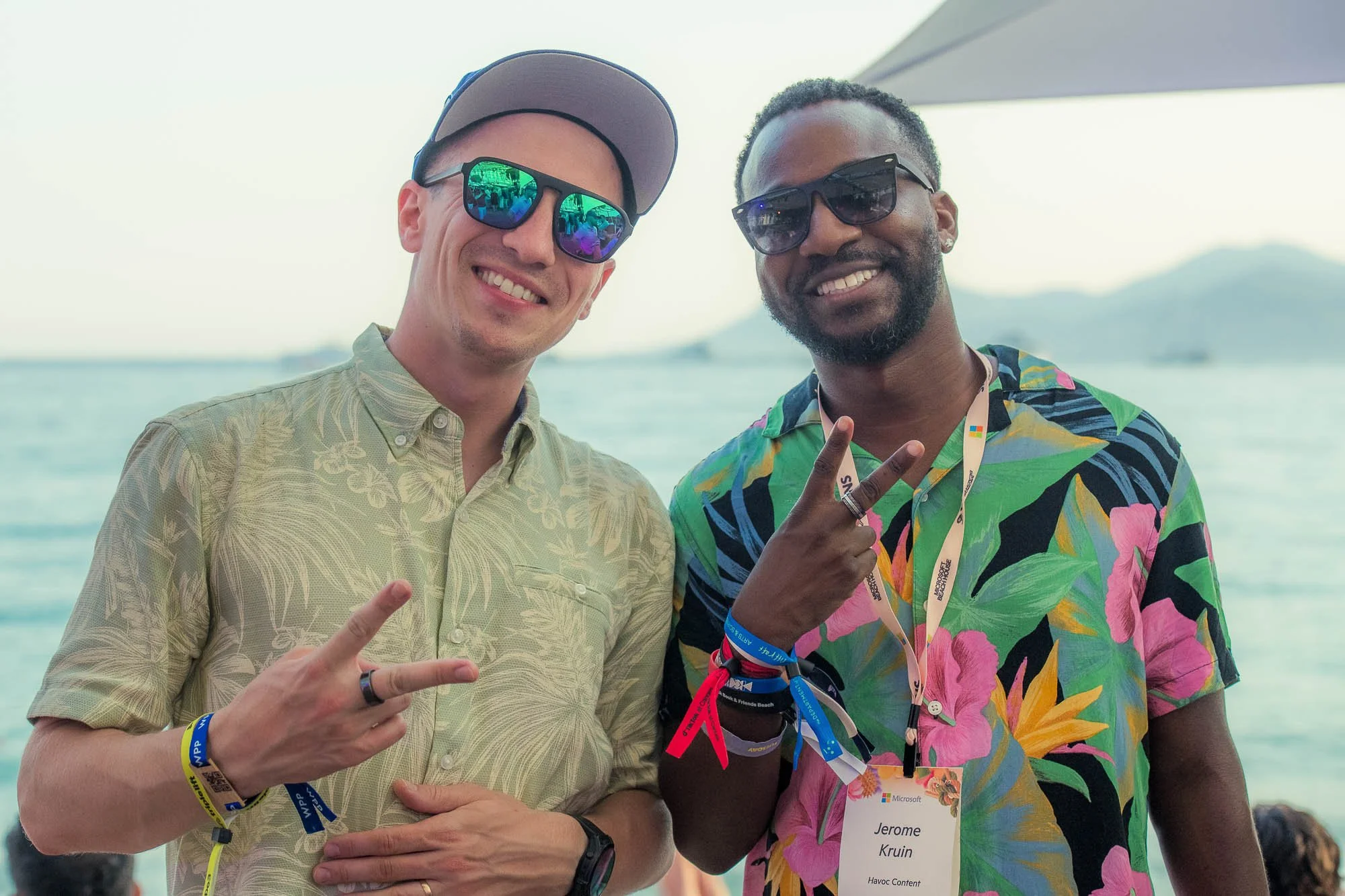 Two men wearing sunglasses and colorful shirts smiling and making peace signs, standing outdoors in front of water and mountains.