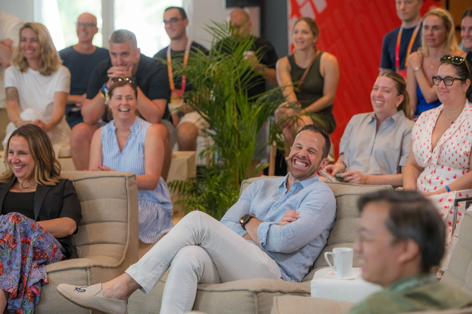 Group of people sitting and standing in a bright room, laughing and enjoying a presentation or event, with a large green plant in the center and a red patterned wall in the background.