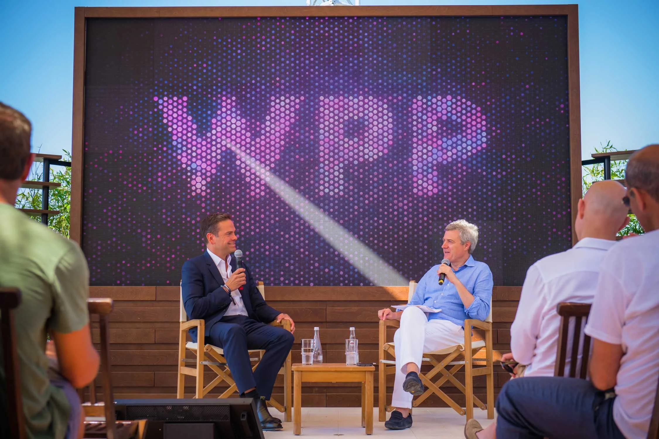 Two men are seated on a stage holding microphones, engaged in a discussion during a live event. Behind them is a large digital screen displaying the word 'VIVO' in pink and purple pixelated letters. Several audience members are seated in front, liste