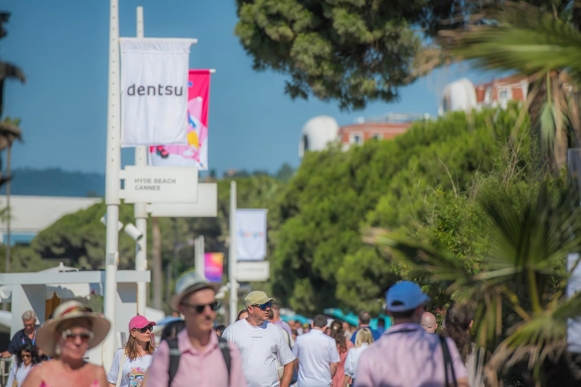 A crowd of people walking outdoors on a sunny day with trees and park signs, including one that says 'Hyde Beach Cannes' and banners that say 'dentsu'.