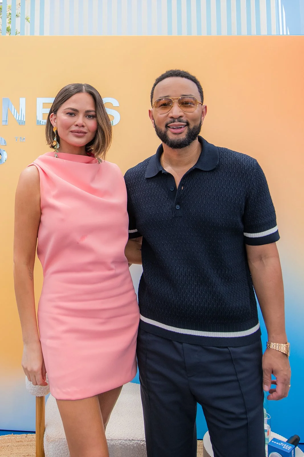 A woman in a pink dress standing next to a man in a dark polo shirt, both smiling at a daytime outdoor event with a yellow backdrop.
