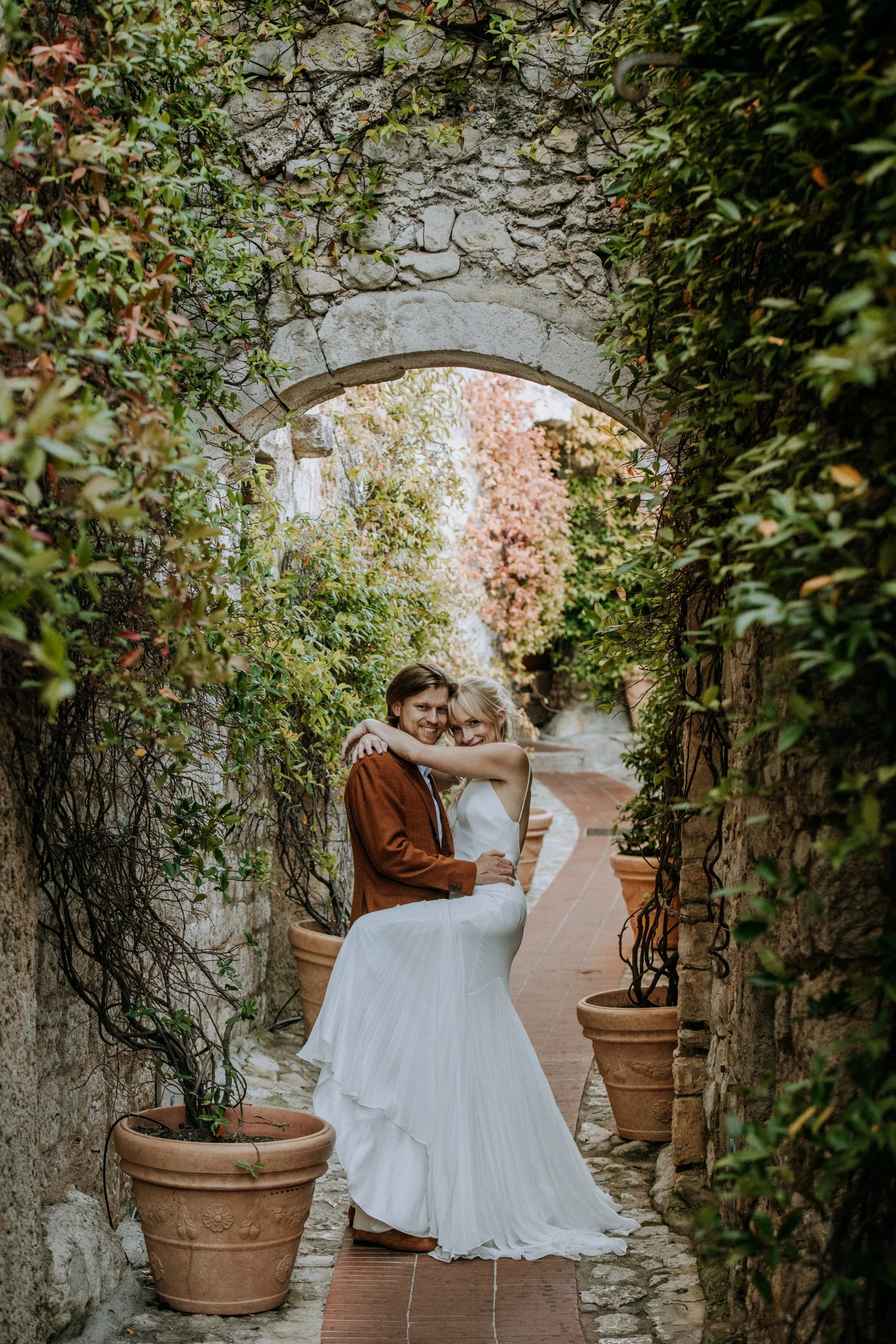 A woman in a white dress is being lifted by a man in a brown jacket in a narrow, garden pathway, surrounded by potted plants and vines, with an stone arch overhead.