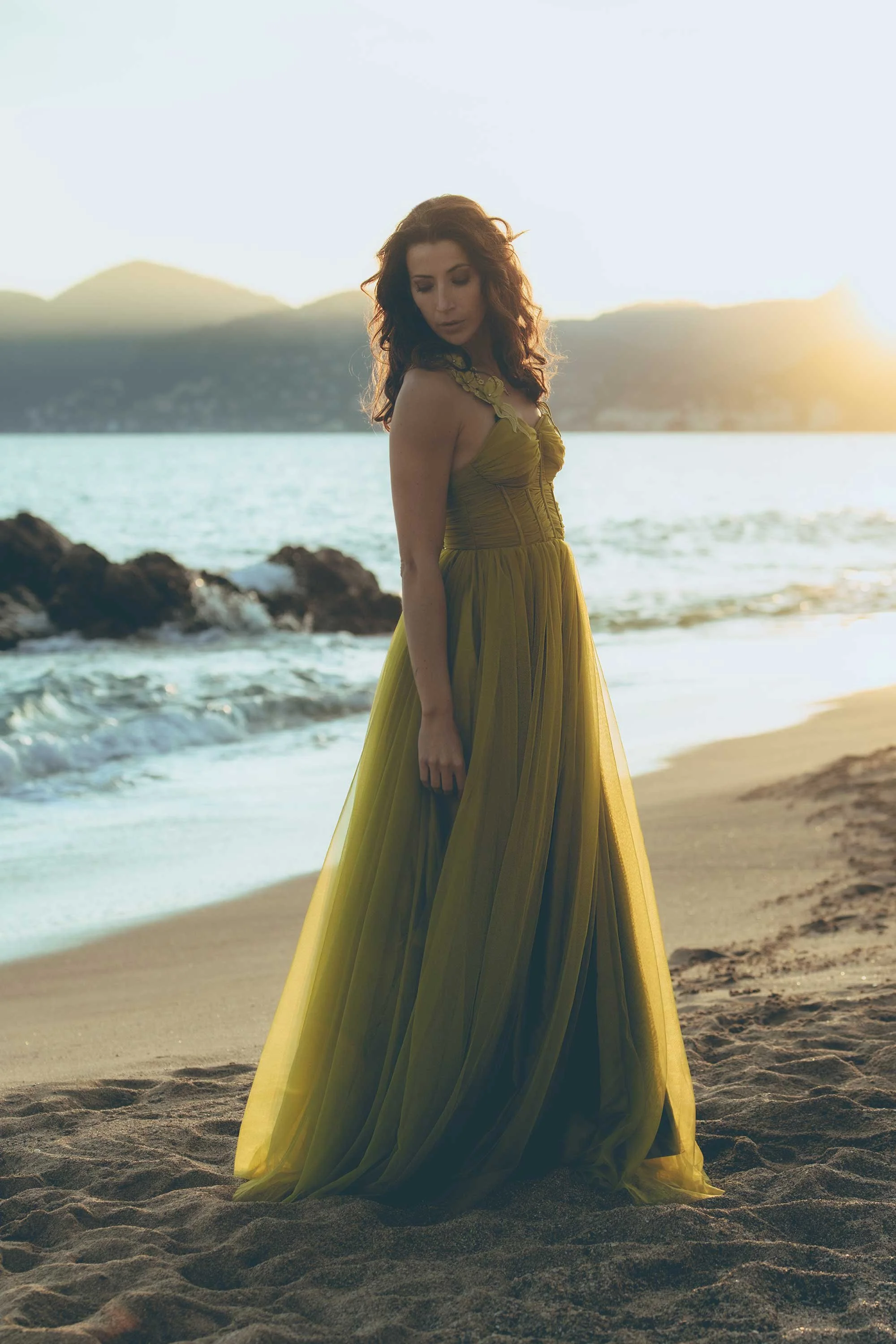 A woman in a long yellow dress stands on a sandy beach during sunset with mountains and water in the background.