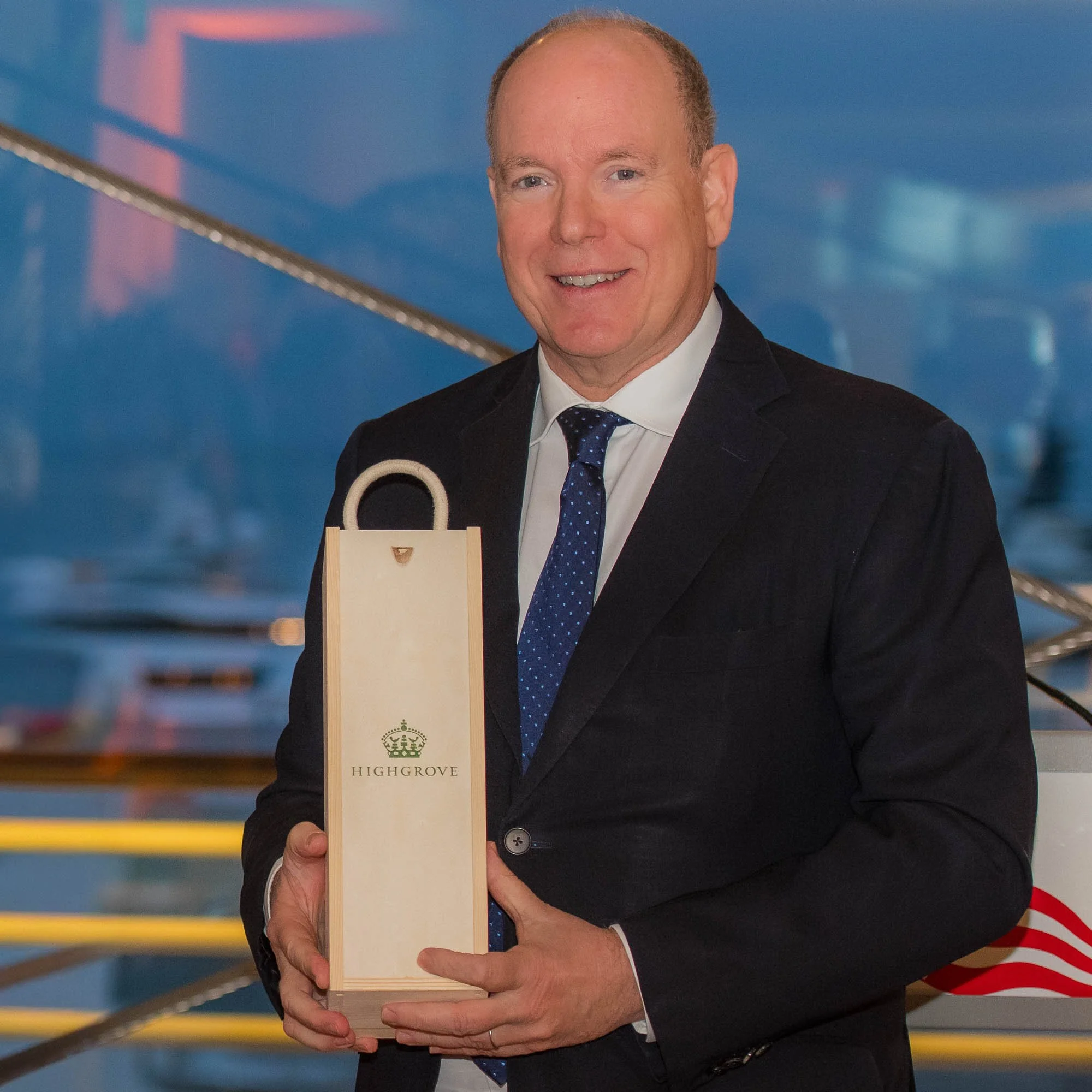 A man in a black suit, white shirt, and blue tie holding a wooden box with the Highgrove logo and crown. The background shows a blue mural with a red and white striped object partially visible.