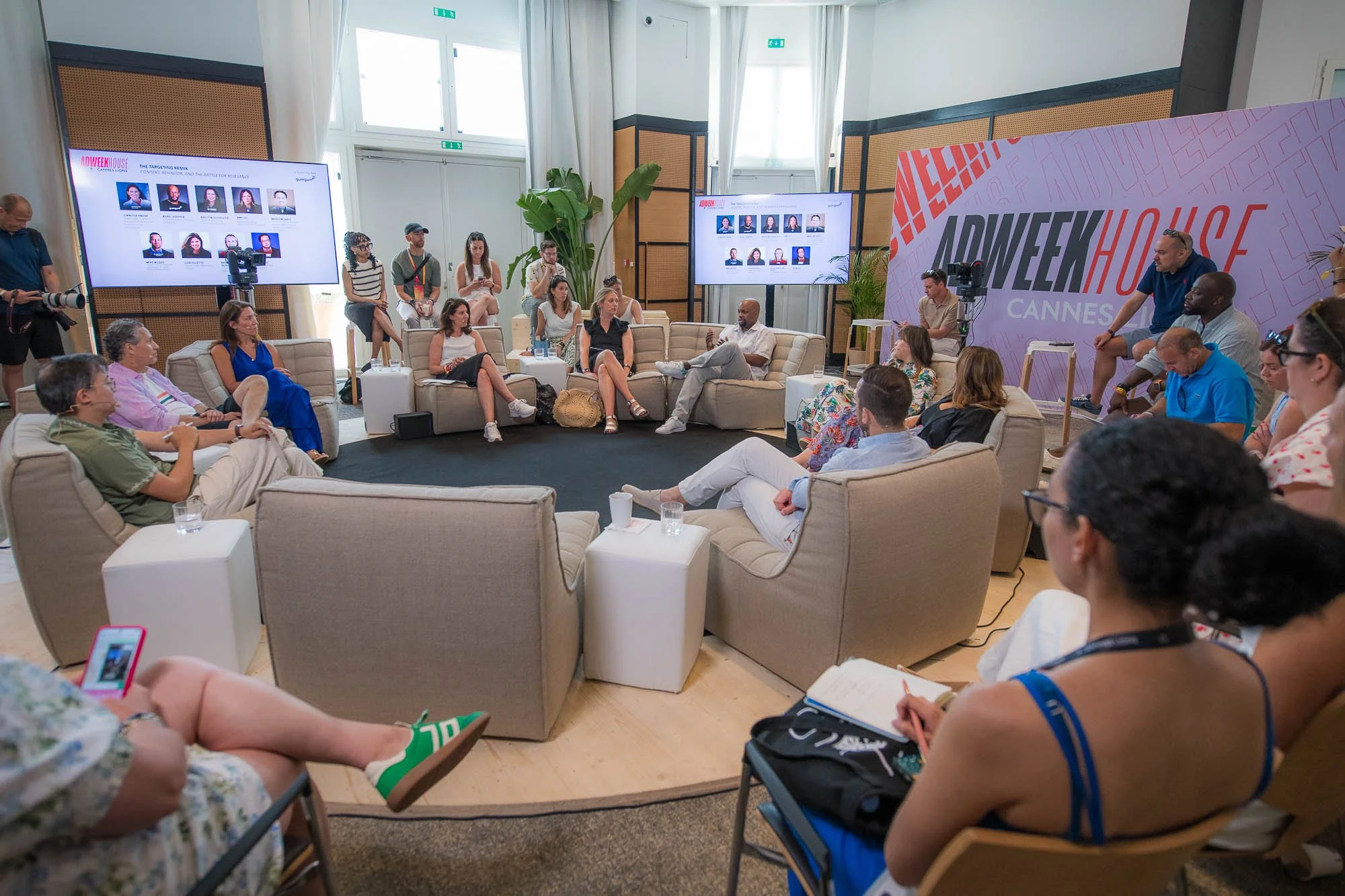 A panel discussion at the Cannes Adweek House event, with people seated in a semi-circle and two large screens displaying photographs of speakers.