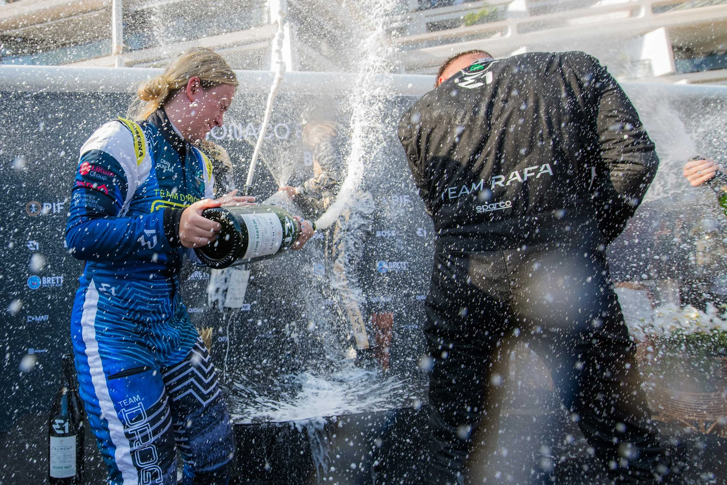 A woman in a blue racing suit is celebrating by spraying champagne, with a bottle in her hand, in front of a backdrop with logos, while another person dressed in black stands beside her at an outdoor event.