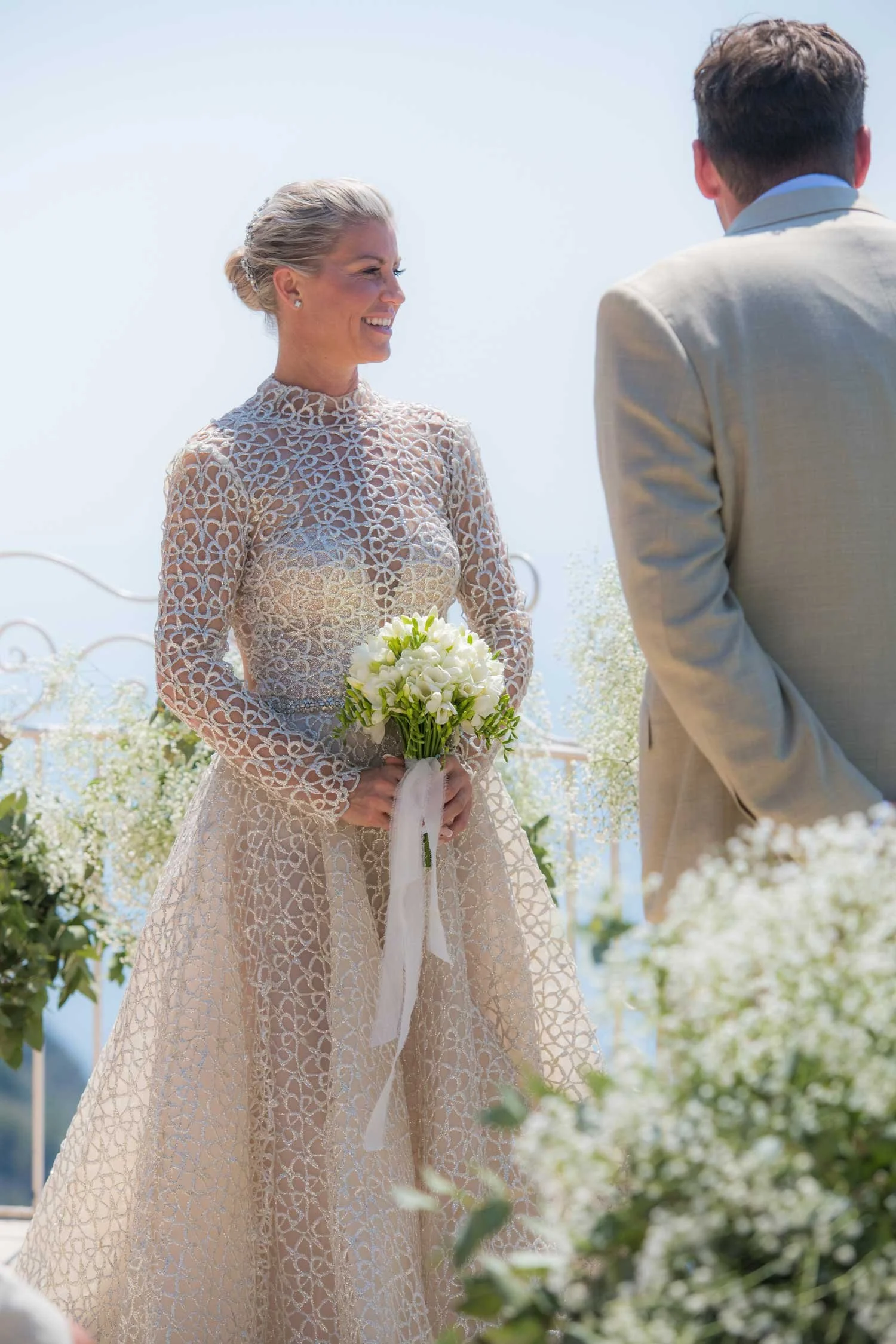 Bride in a sheer, patterned wedding dress holding a bouquet of white flowers during an outdoor wedding ceremony.