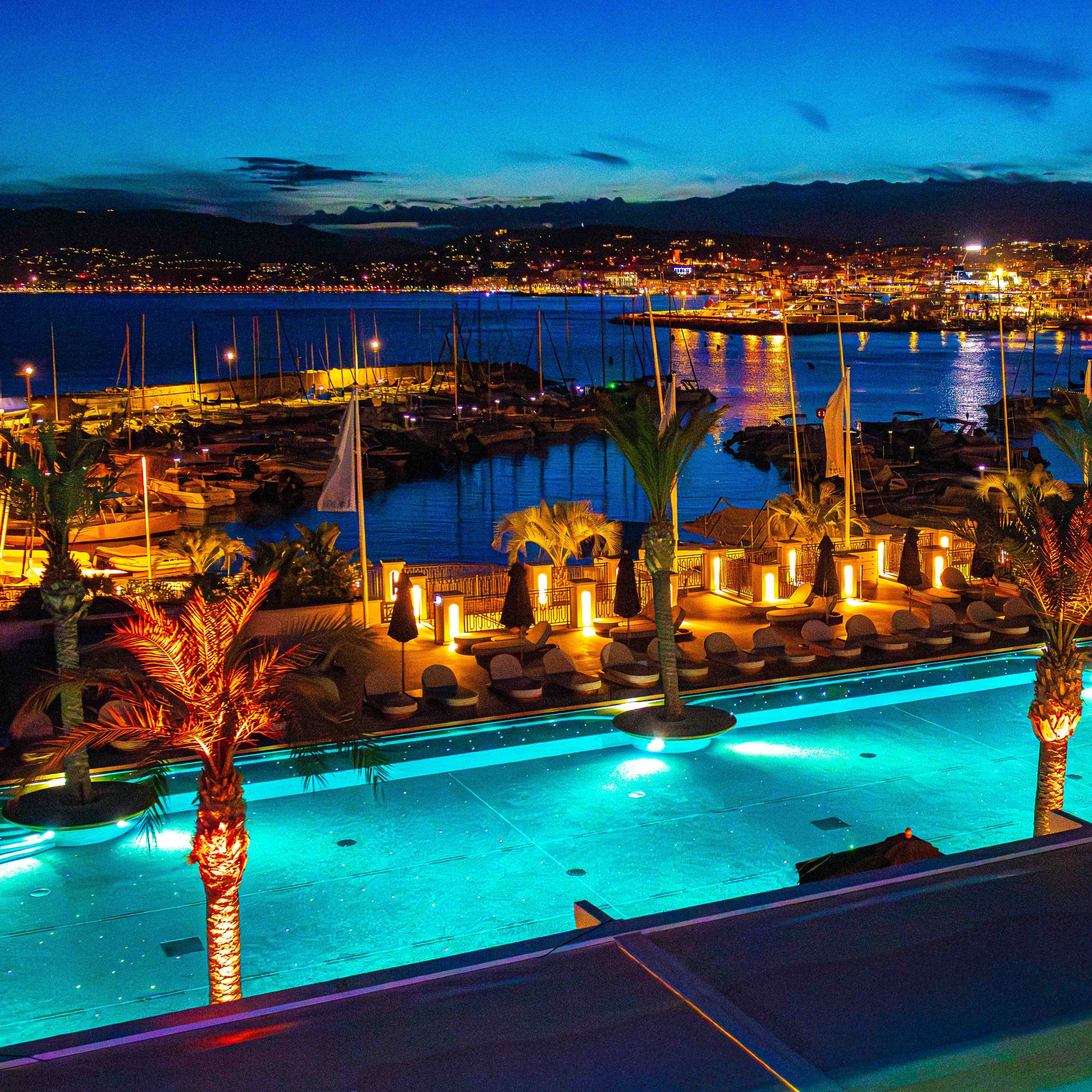 A night view of a marina with boats docked in calm water, illuminated by colorful lights, and a cityscape with buildings and mountains in the background.