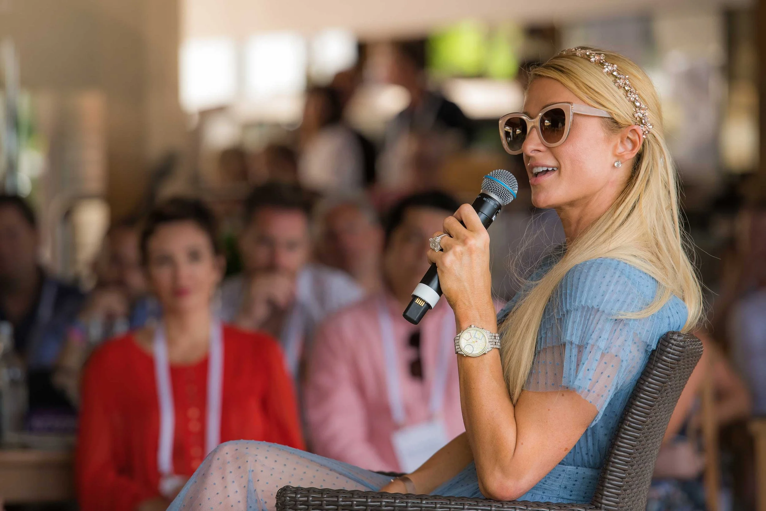 A woman with long blonde hair, wearing sunglasses, a headband, and a blue dress, holds a microphone while speaking to an audience in a bright indoor setting.
