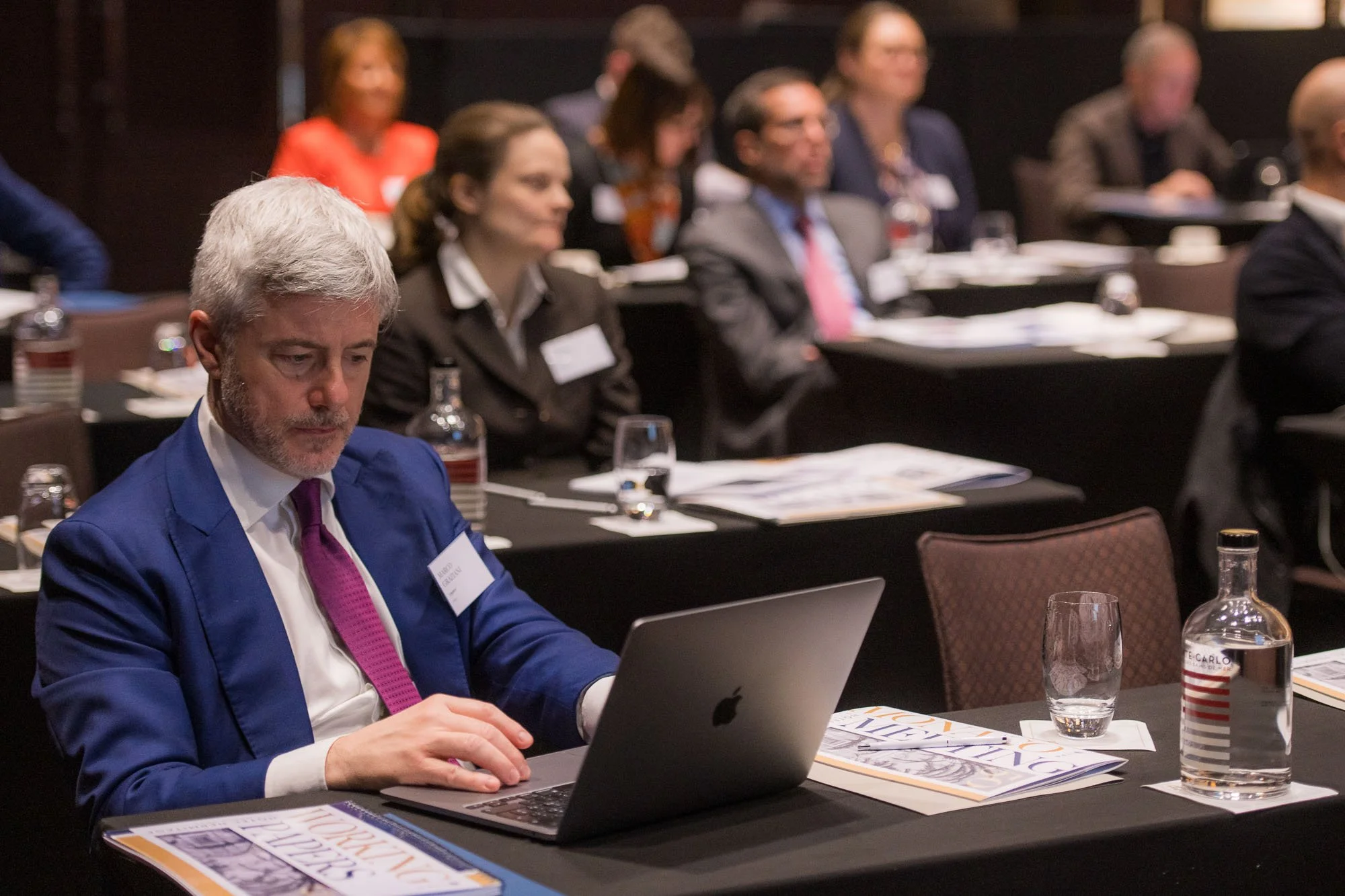 A man in a blue suit and pink tie working on a laptop at a conference table with several other attendees seated behind him, papers, water bottles, and glasses on the table.