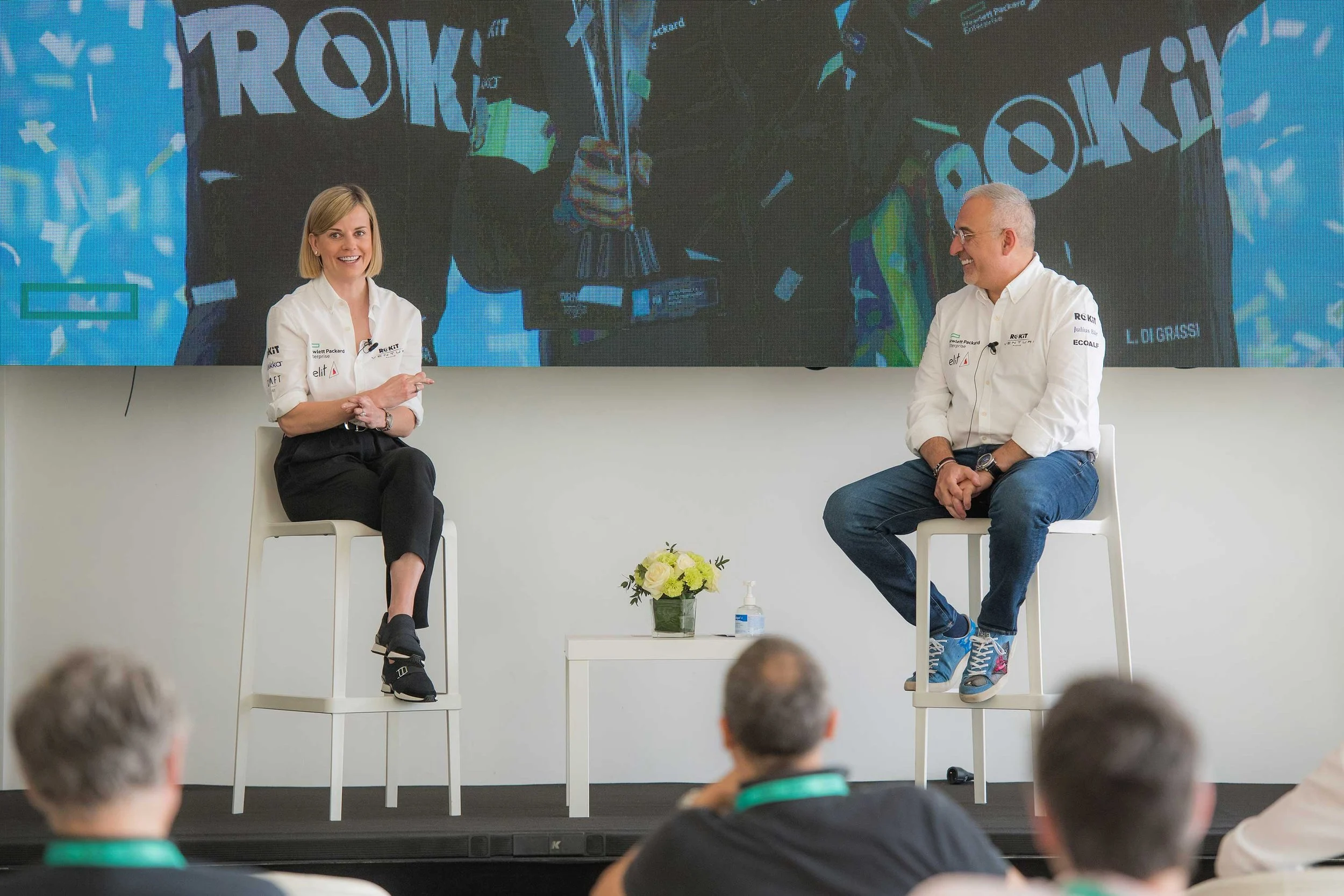Two people sitting on chairs on a stage in front of a large digital screen, with an audience watching. The woman on the left has blonde hair, is smiling, and is wearing a white shirt and black pants. The man on the right has gray hair, is smiling, and is wearing glasses, a white shirt, and blue jeans. There is a small table with a flower arrangement and hand sanitizer in front of them.